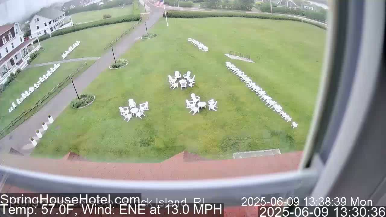A grassy lawn with numerous white chairs arranged in rows and small groups around tables, viewed from above in windy conditions.