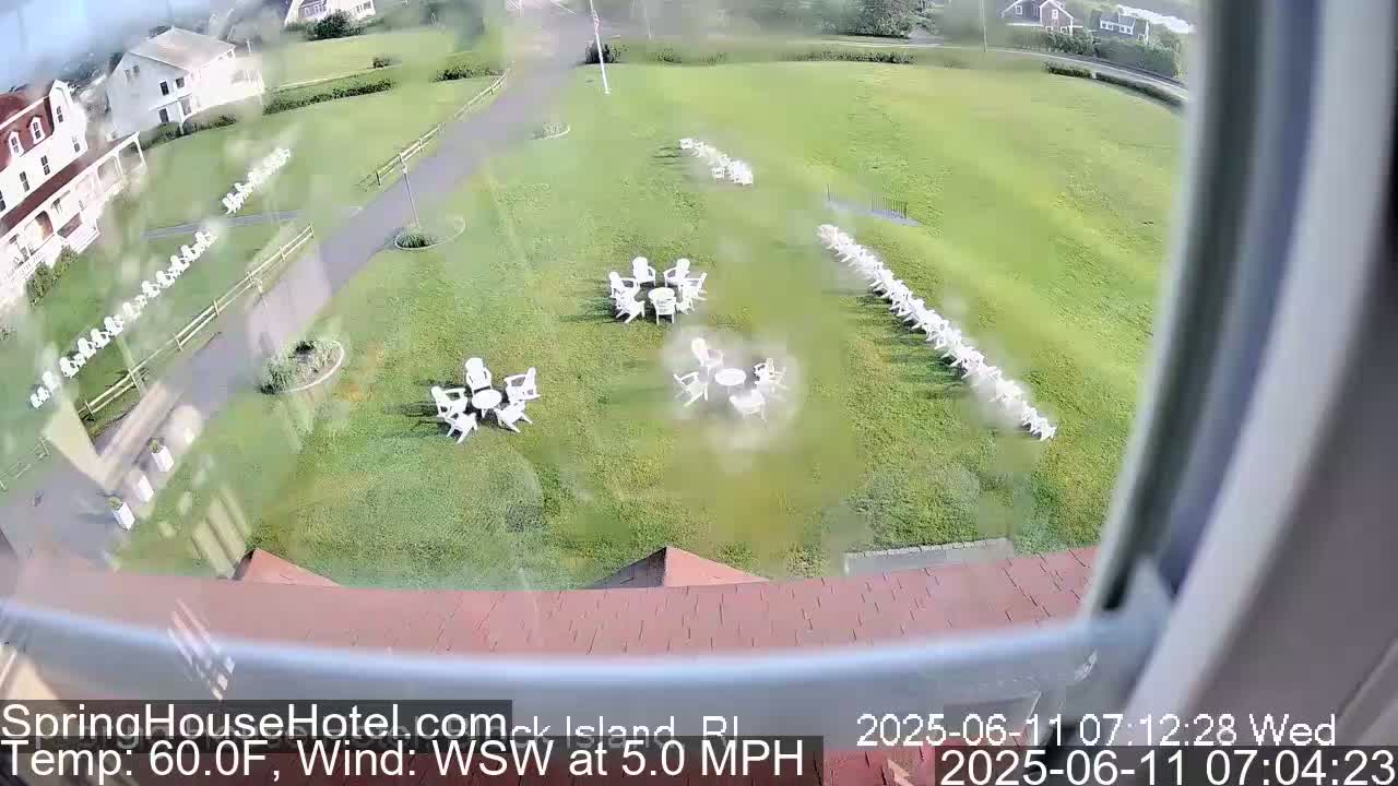 An elevated view shows a grassy area with numerous white chairs arranged in rows and small clusters around tables, overlooking a building and under a partly cloudy sky with a gentle wind.