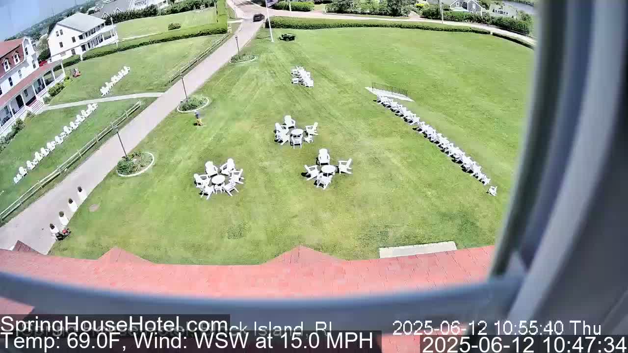 A high-angle view shows a grassy area with numerous white chairs arranged in rows and small clusters around tables, next to a building and a paved path, on a sunny day with a wind of 15 mph from the west-southwest.