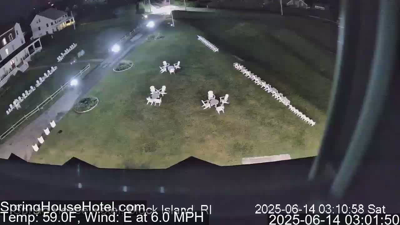 A nighttime aerial view shows a grassy area with numerous white chairs arranged in rows and small groups around tables, near a building and a paved walkway, under a clear sky with an east wind.