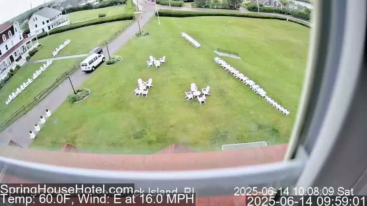 A white van is driving on a paved path next to a grassy area with numerous white lawn chairs and small tables arranged in groups, under an overcast sky with a 16 mph east wind and a temperature of 60°F.