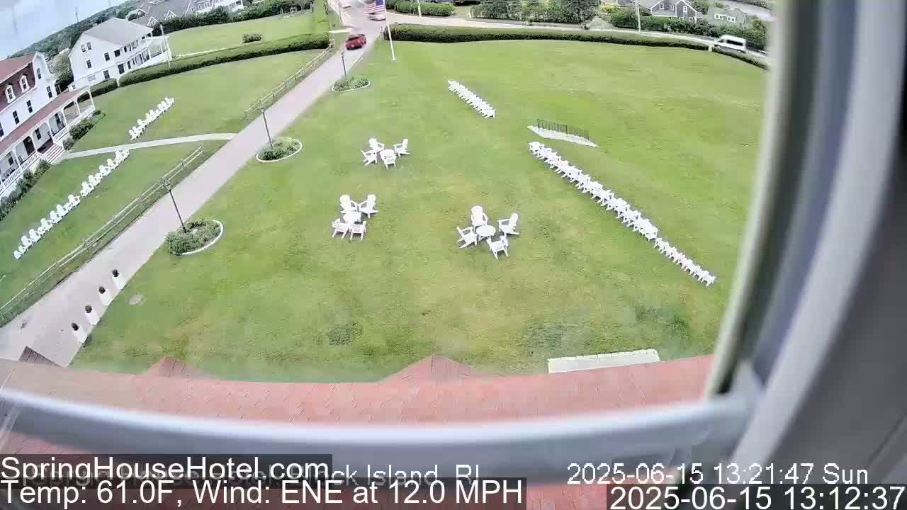 A sunny, outdoor scene shows a large grassy area with numerous white chairs arranged in rows and small groups around tables, situated near a building and a paved path.