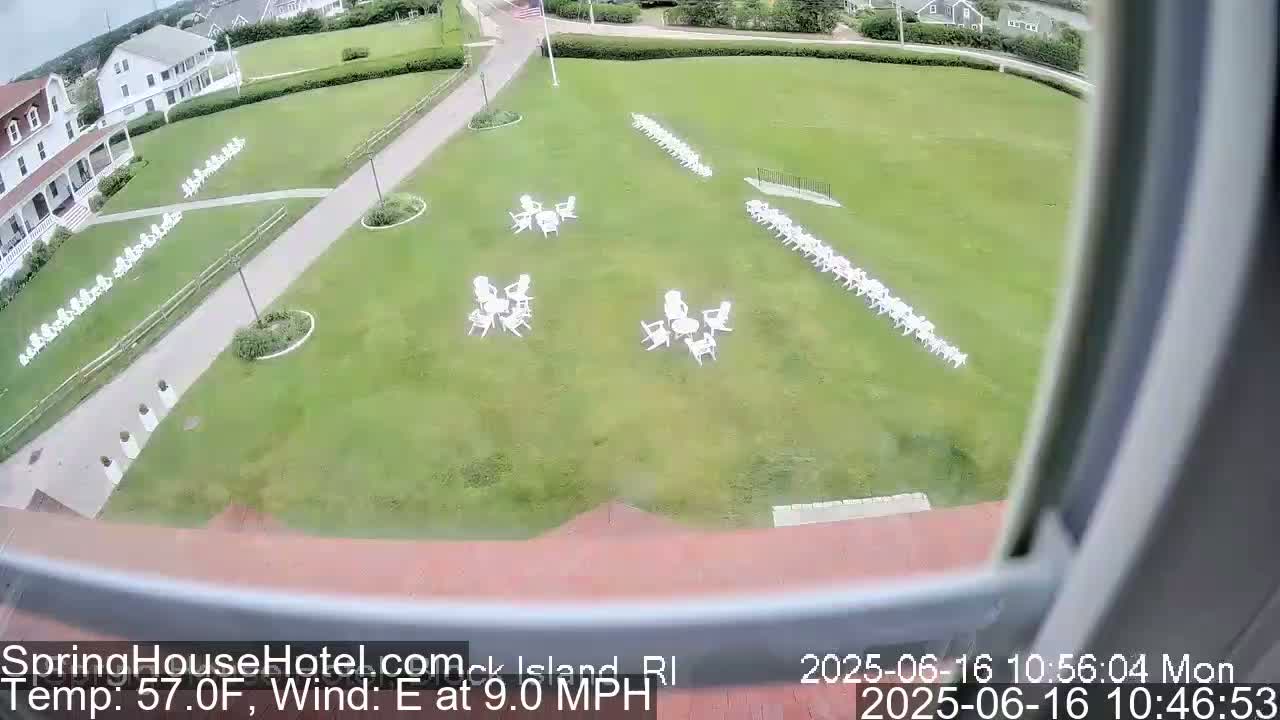 A grassy lawn with numerous white chairs arranged in rows and small groups of tables and chairs, viewed from an elevated position on a partly sunny day with an east wind at 9 mph.