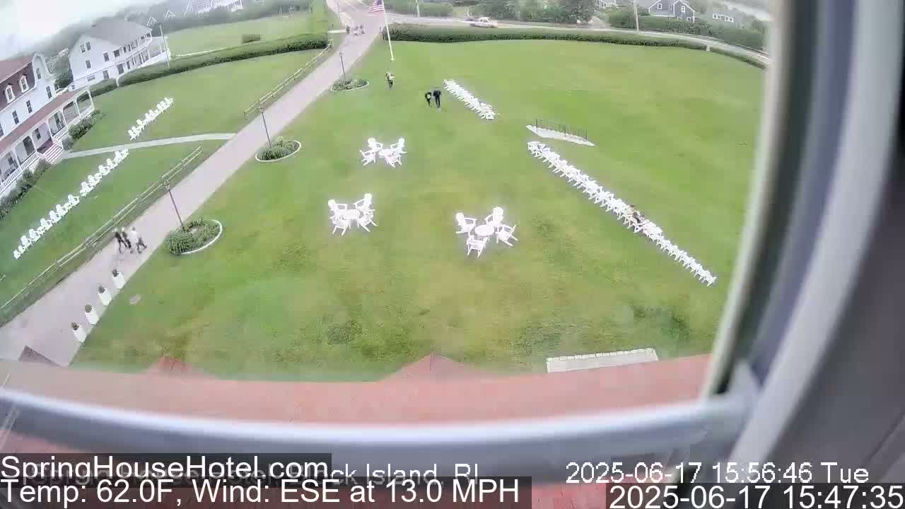 A grassy lawn with white chairs arranged in rows and small groupings of tables and chairs, viewed from above on a partly cloudy day with a 13 mph wind from the east-southeast.