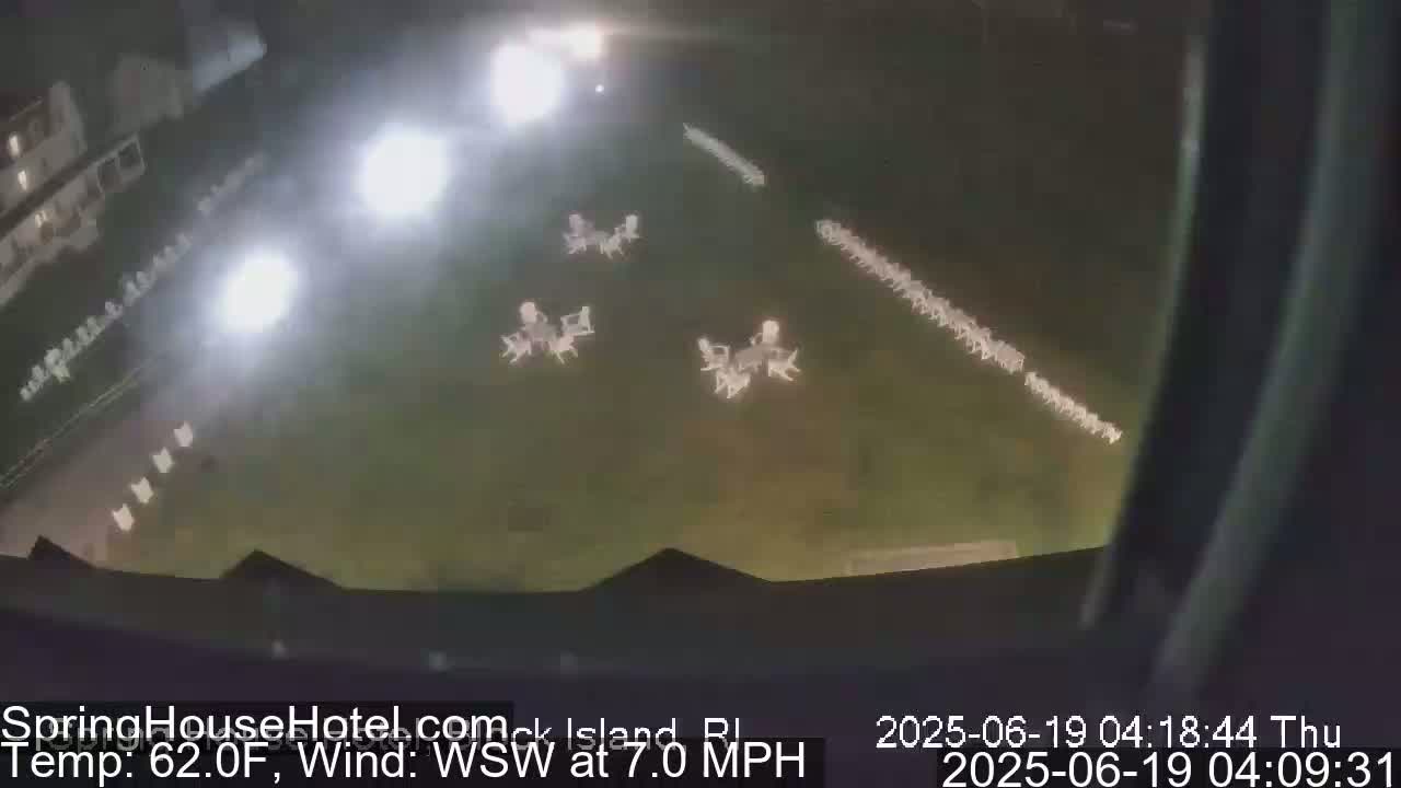 A nighttime aerial view shows a grassy area with several sets of white patio furniture arranged in rows and small clusters, illuminated by bright lights.