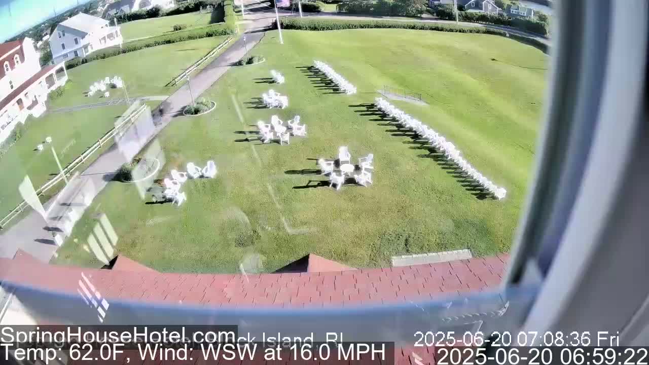 A grassy area with numerous white chairs arranged in groups and rows is seen from a window, with a building and a walkway visible in the background, under sunny conditions with a west-southwest wind at 16 mph.