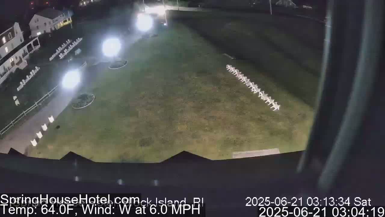 An aerial nighttime view shows a grassy area with a line of chairs, illuminated by bright lights, adjacent to a building.