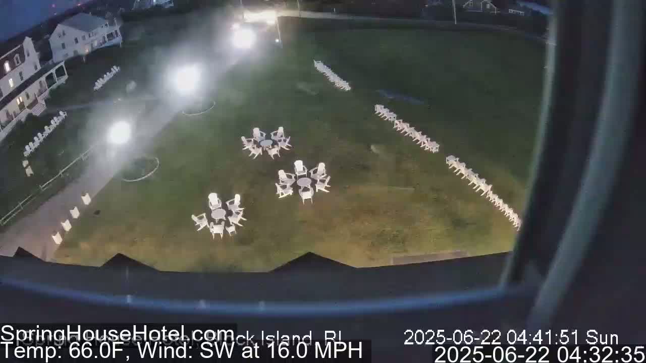 An aerial nighttime view shows a grassy area with several sets of white chairs and tables arranged around it, near a building, under a partly cloudy sky with a southwest wind at 16 mph.