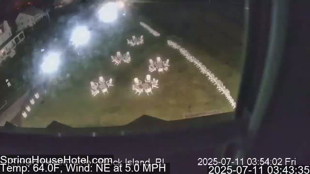 A nighttime aerial view shows a grassy area with numerous white tables and chairs arranged in rows and small clusters, illuminated by bright lights.
