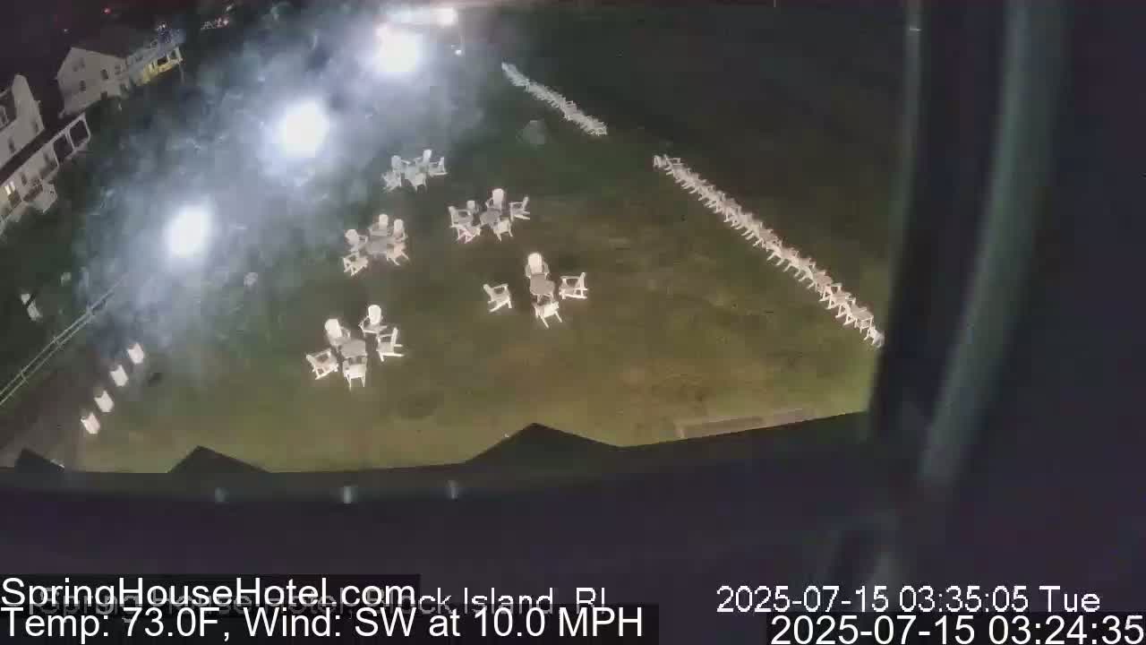 An outdoor nighttime scene shows numerous empty white chairs arranged in rows and small groups across a grassy area, illuminated by bright lights, with a southwest wind at 10 mph and a temperature of 73 degrees Fahrenheit.