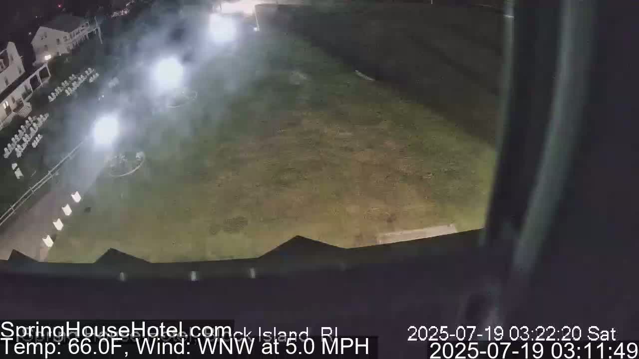 A nighttime, high-angle view shows a grassy area with bright lights and some hazy fog, adjacent to a building with white chairs arranged on its grounds, under a WNW wind at 5.0 mph and a temperature of 66.0°F.