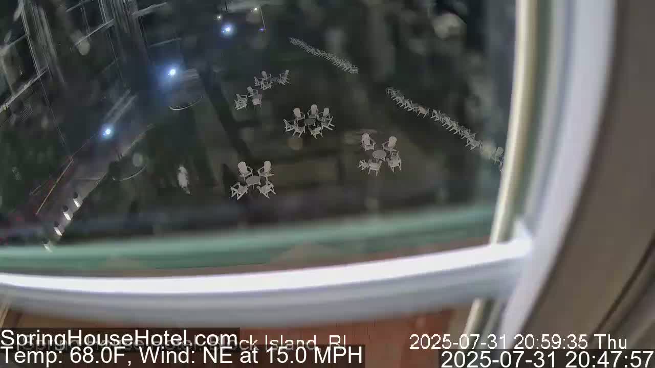 A nighttime view through a window shows a patio area with numerous empty white chairs and tables arranged in groups, with a northeast wind at 15 mph and a temperature of 68 degrees Fahrenheit.