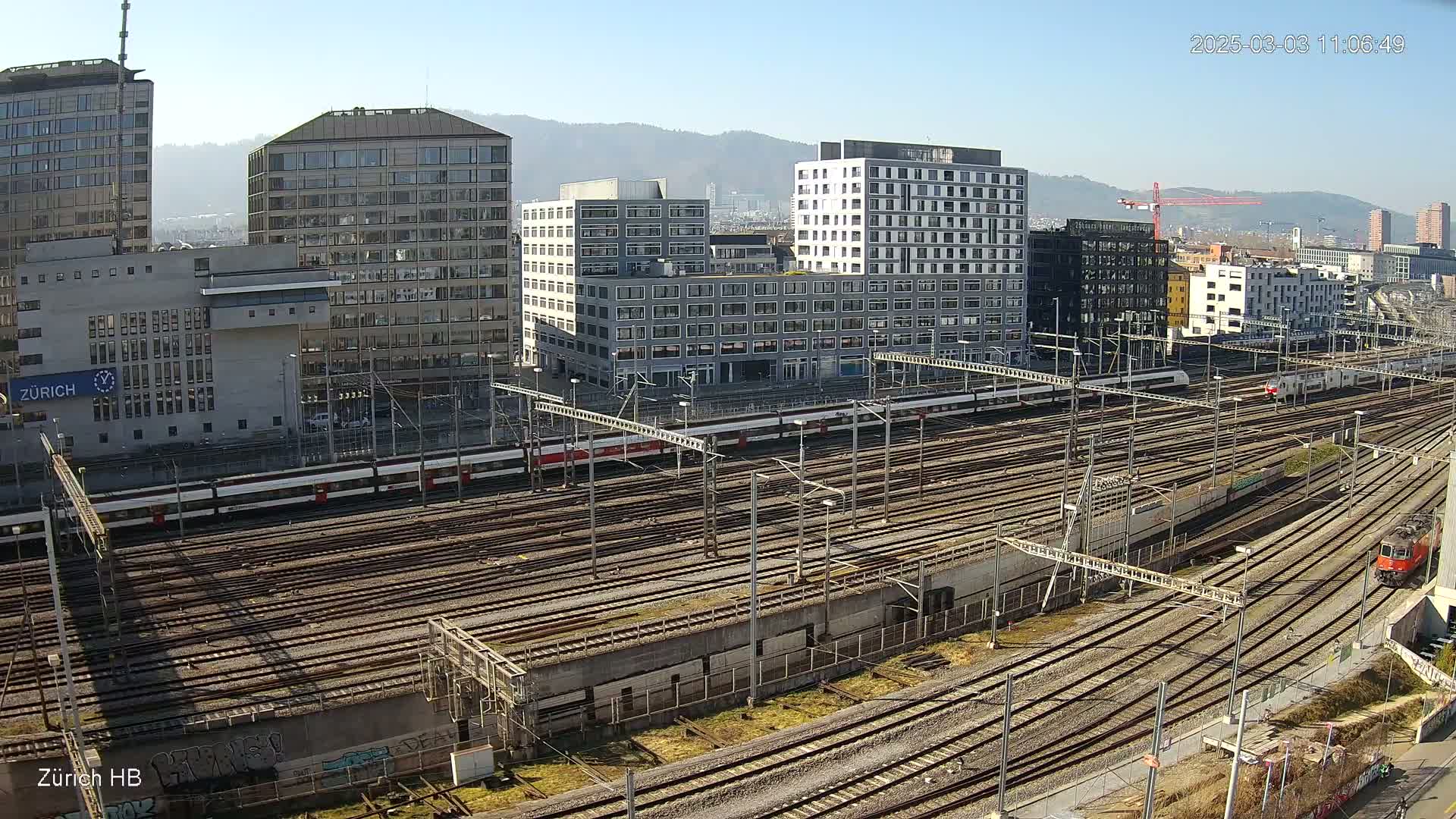 A sunny day overlooking a large railway yard with multiple train tracks, several modern buildings, and a mountain range in the distance.