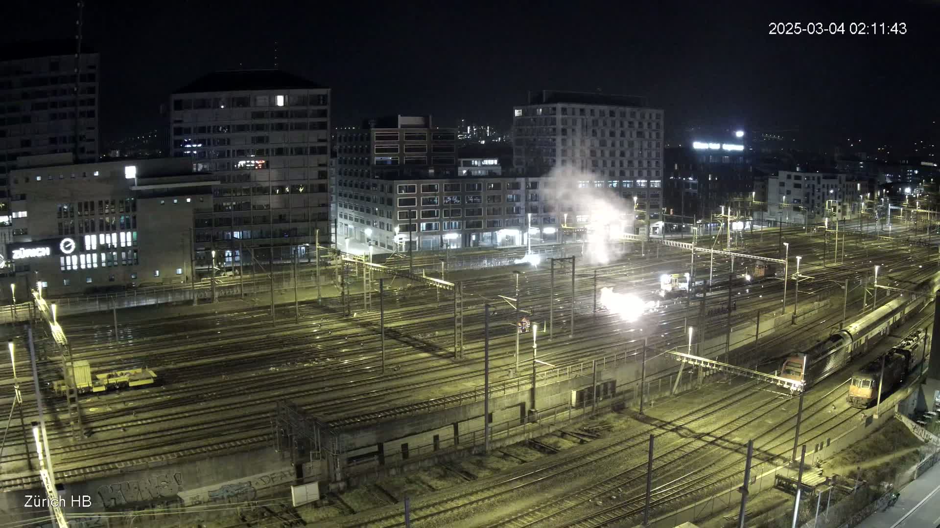 A nighttime, high-angle view of a train yard with multiple train cars and sparks emanating from near one of them, surrounded by city buildings under a dark sky.