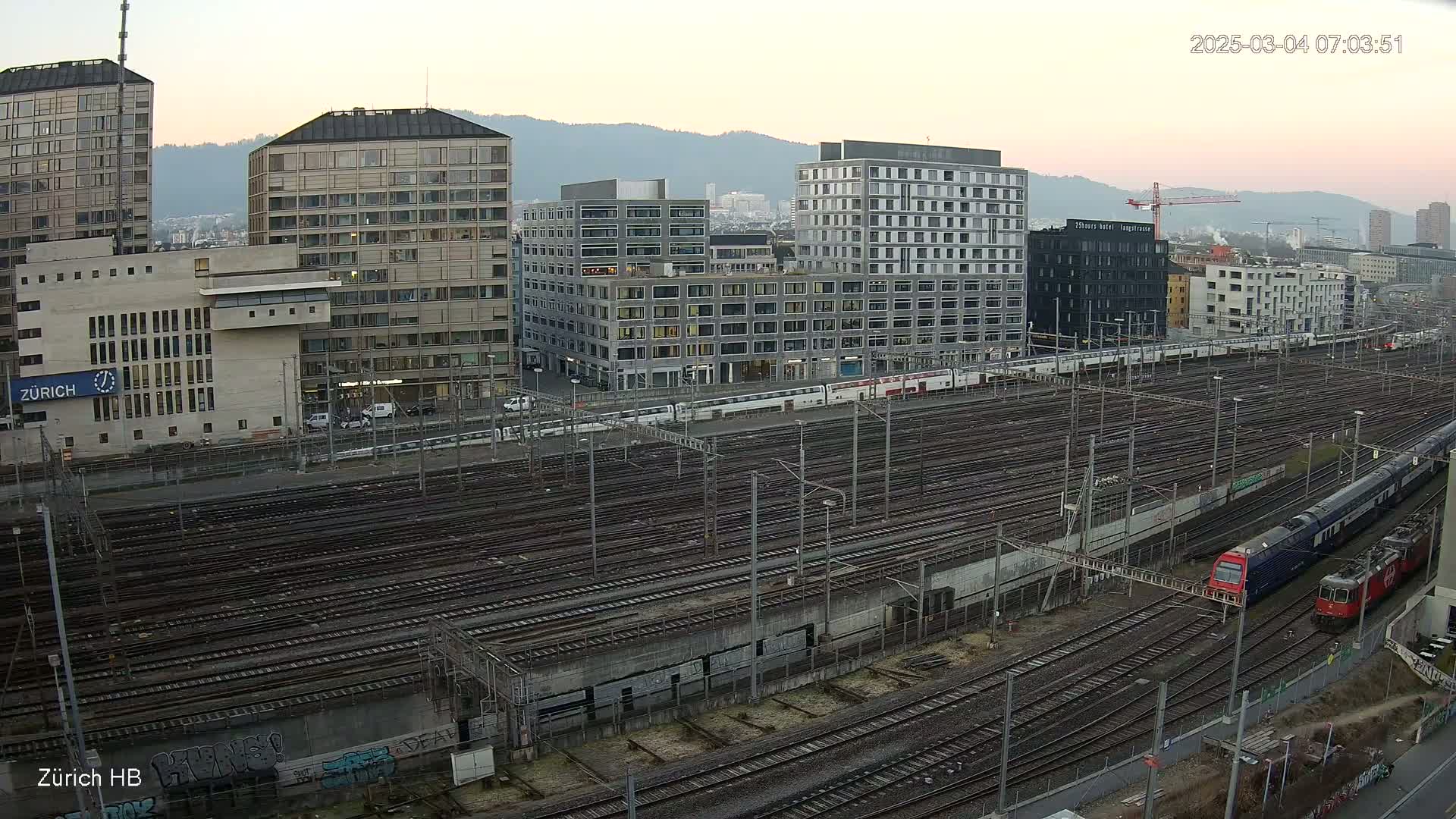 An expansive train yard with numerous tracks, modern buildings, and two trains, all under a twilight sky.