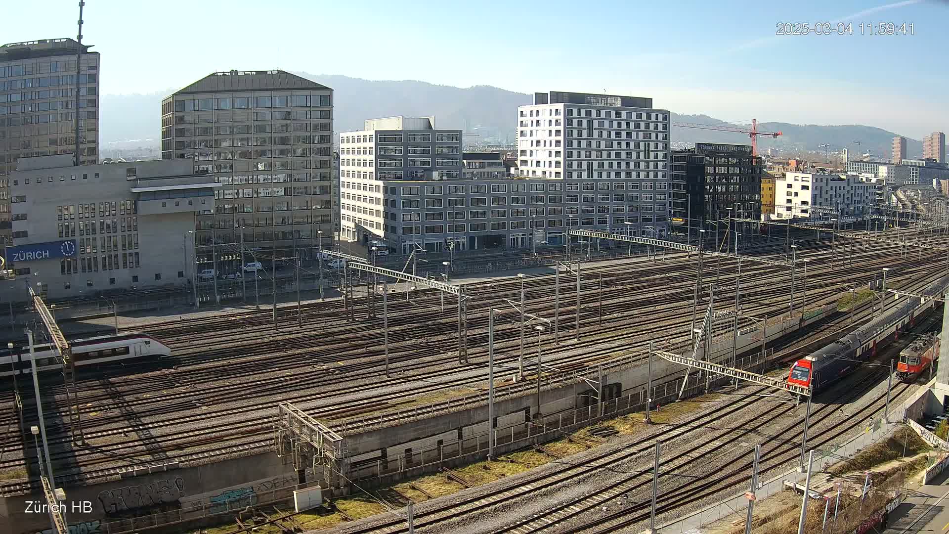 A sprawling railway yard with numerous train tracks and several modern buildings under a clear, sunny sky.
