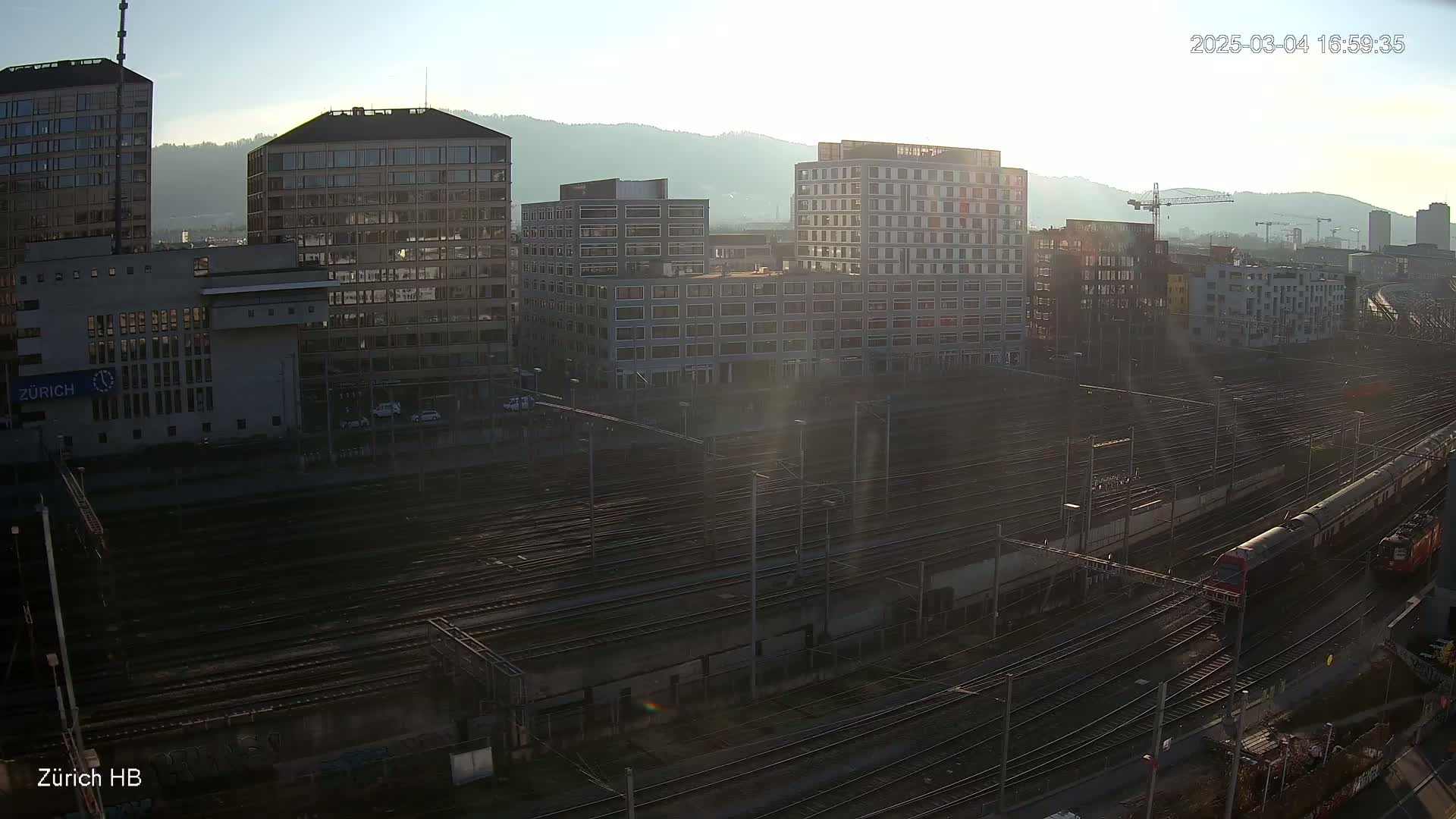 A sprawling train yard with numerous tracks and modern buildings in the background under a bright, sunny sky.