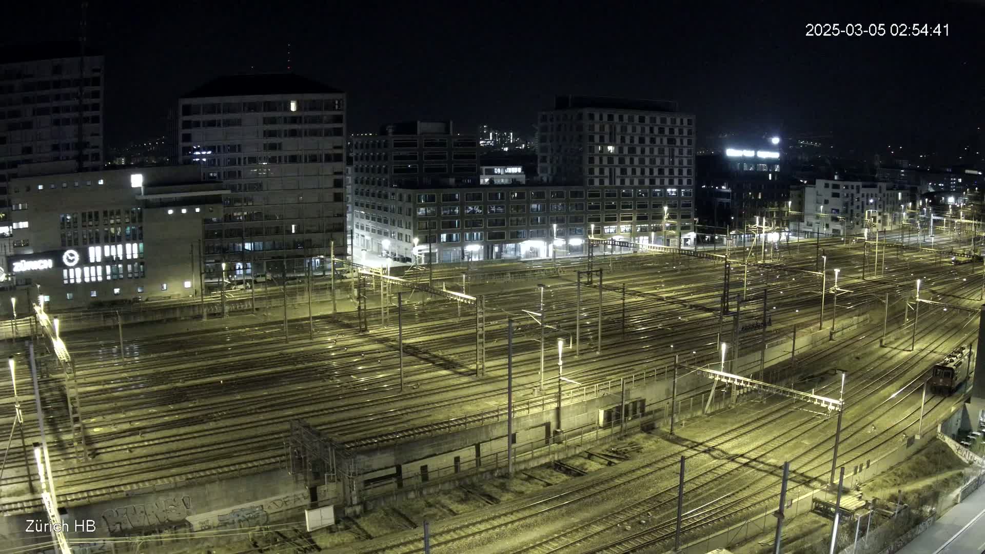 A nighttime aerial view of a large train yard surrounded by city buildings under a clear sky.
