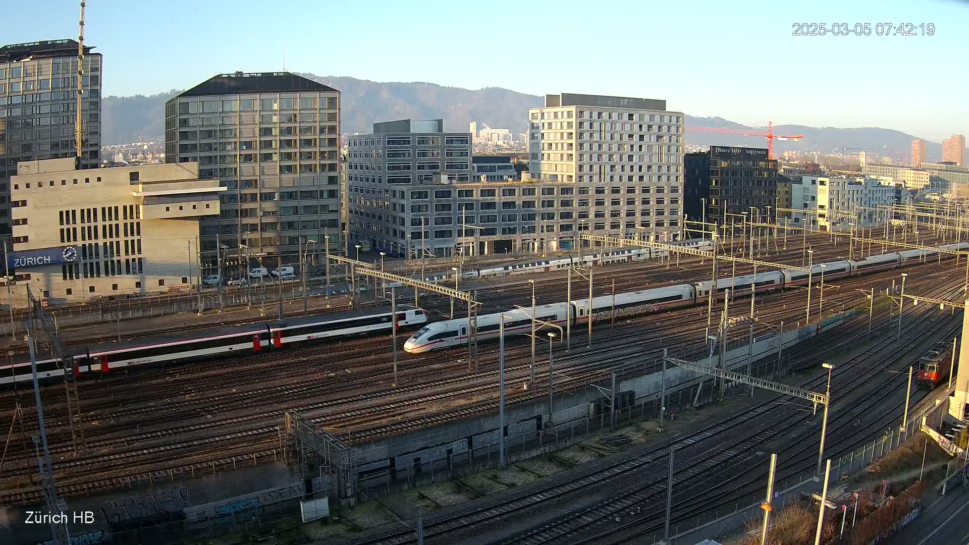 A high-angle view of a train station on a sunny day, showing multiple trains on numerous tracks surrounded by modern buildings and mountains in the distance.