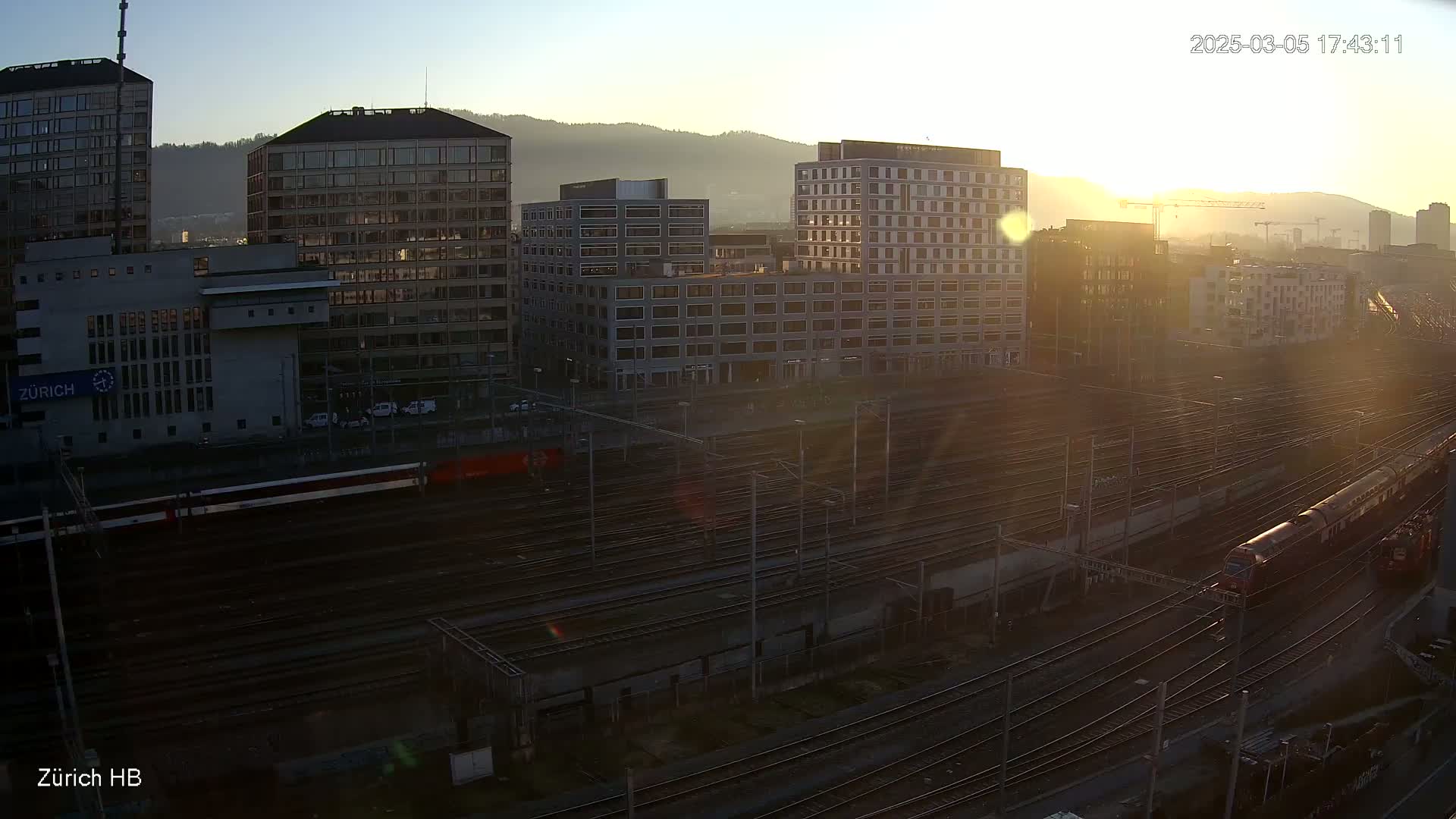 A high-angle, sunlit view of a train station with multiple buildings and many train tracks, during sunrise.