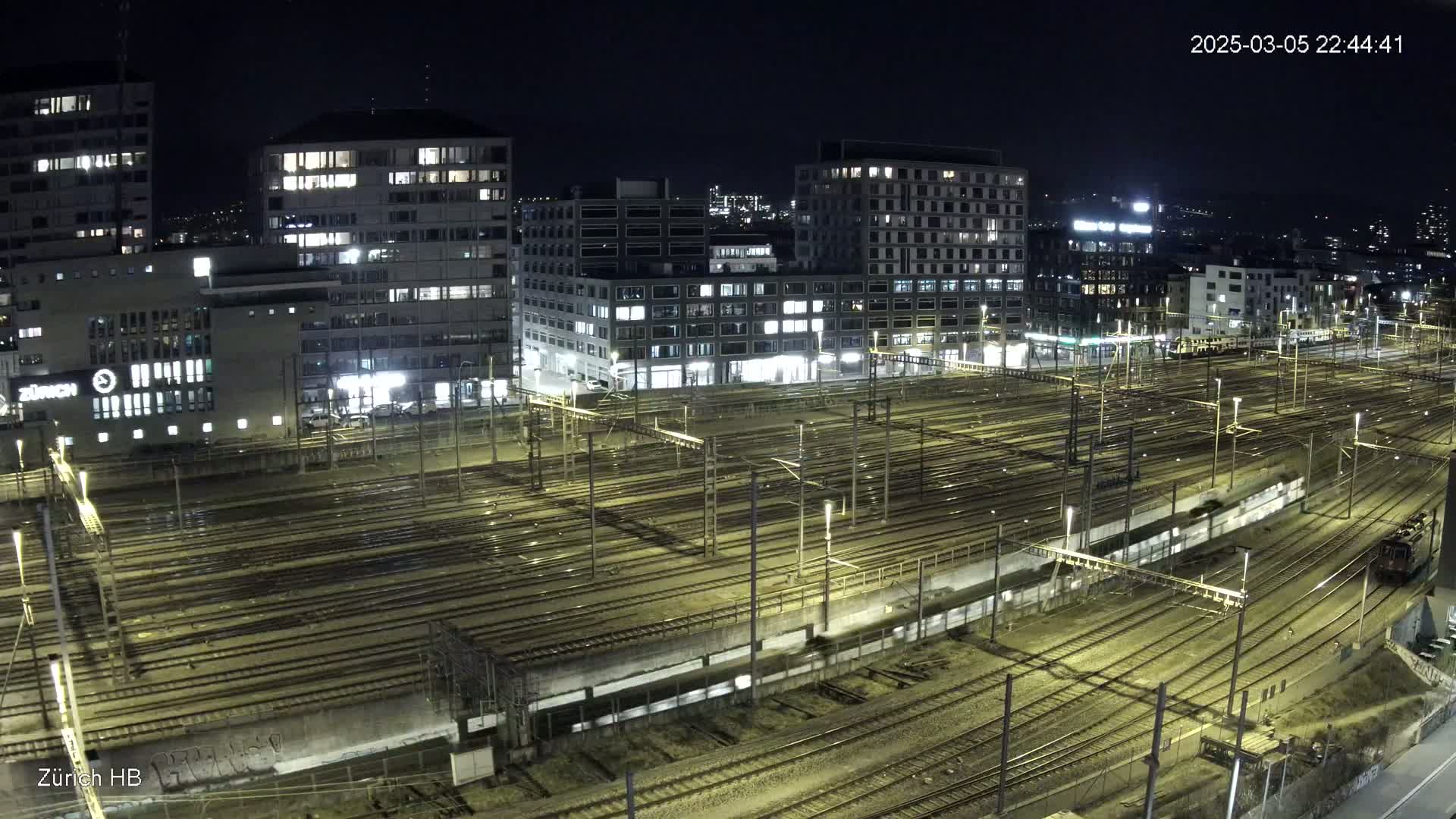 A nighttime aerial view of a large train yard surrounded by illuminated buildings under a clear, dark sky.