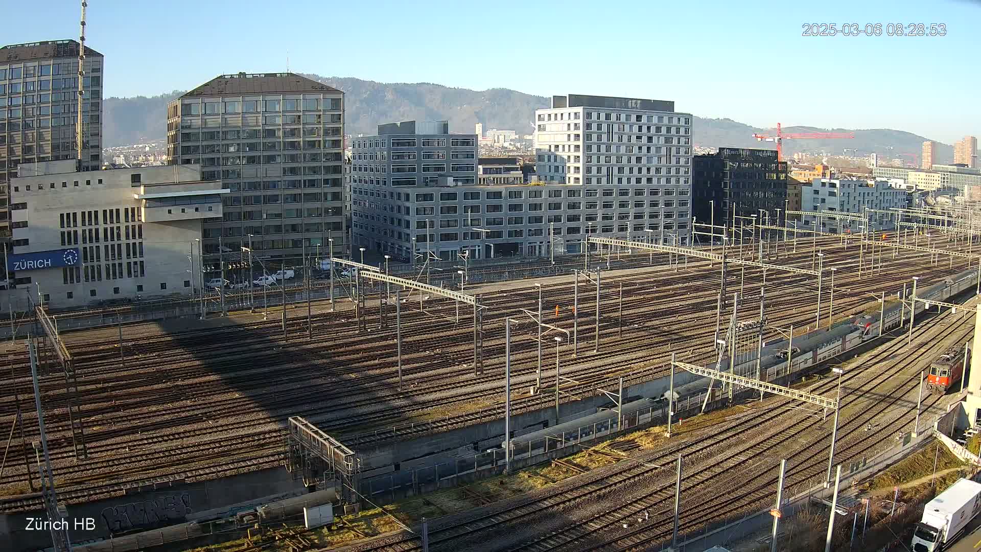 A sprawling railway yard filled with numerous tracks and surrounded by modern buildings under a clear, sunny sky.