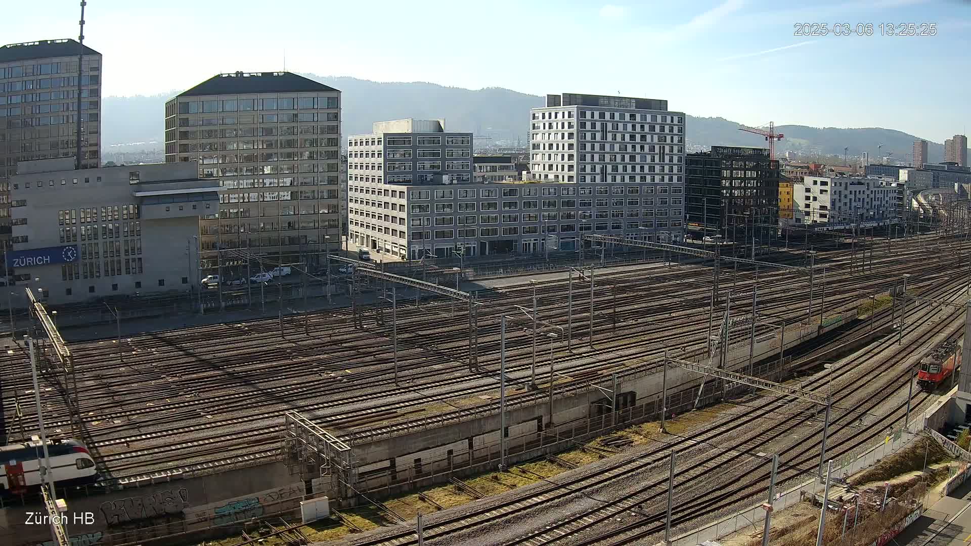 A sunny day reveals a large railway yard surrounded by modern buildings and mountains in the distance.