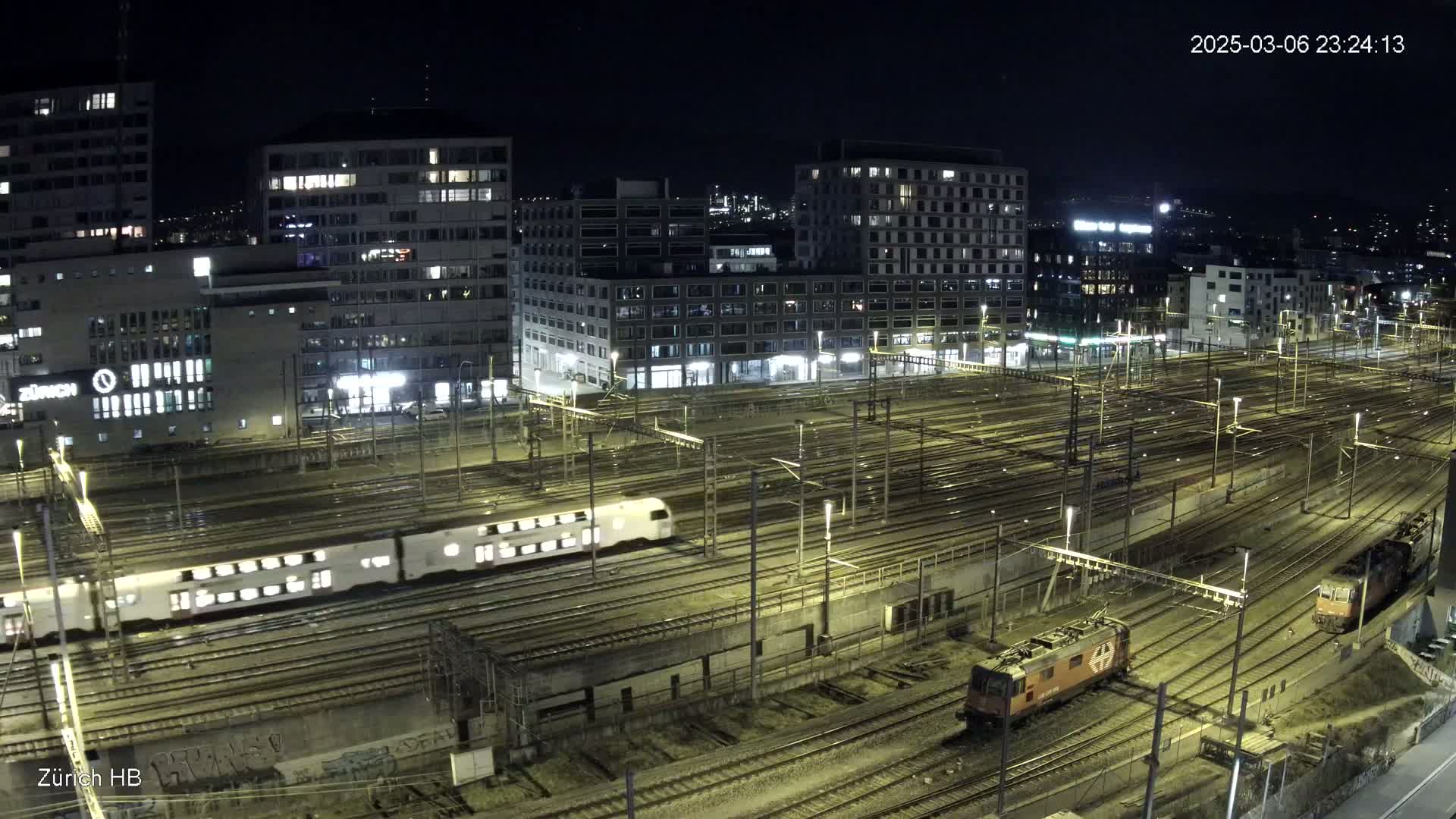 A nighttime aerial view of a train station with multiple tracks, trains, and illuminated buildings surrounding the area.