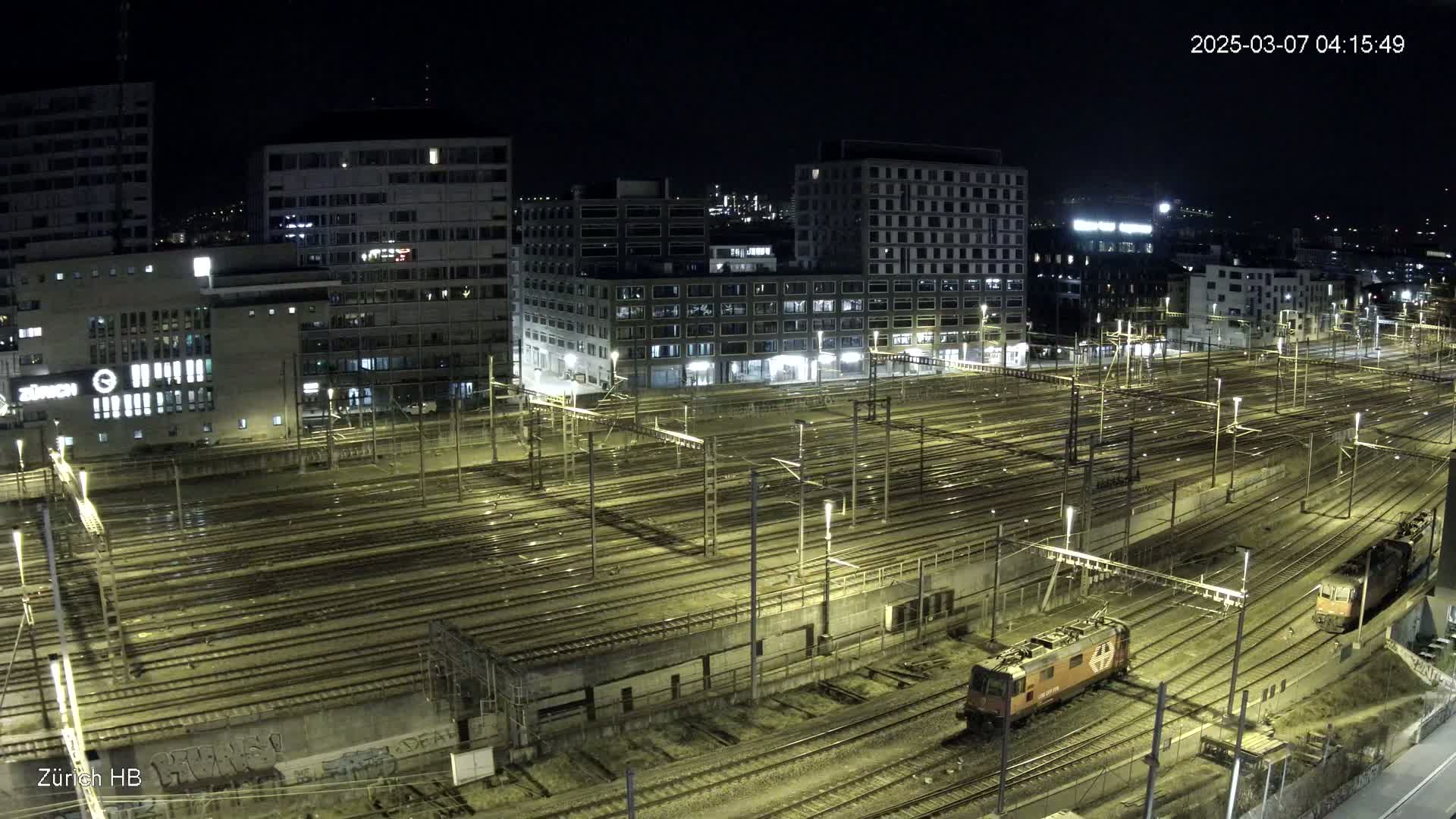A nighttime, high-angle view of a large train yard with numerous tracks and two locomotives, surrounded by city buildings.