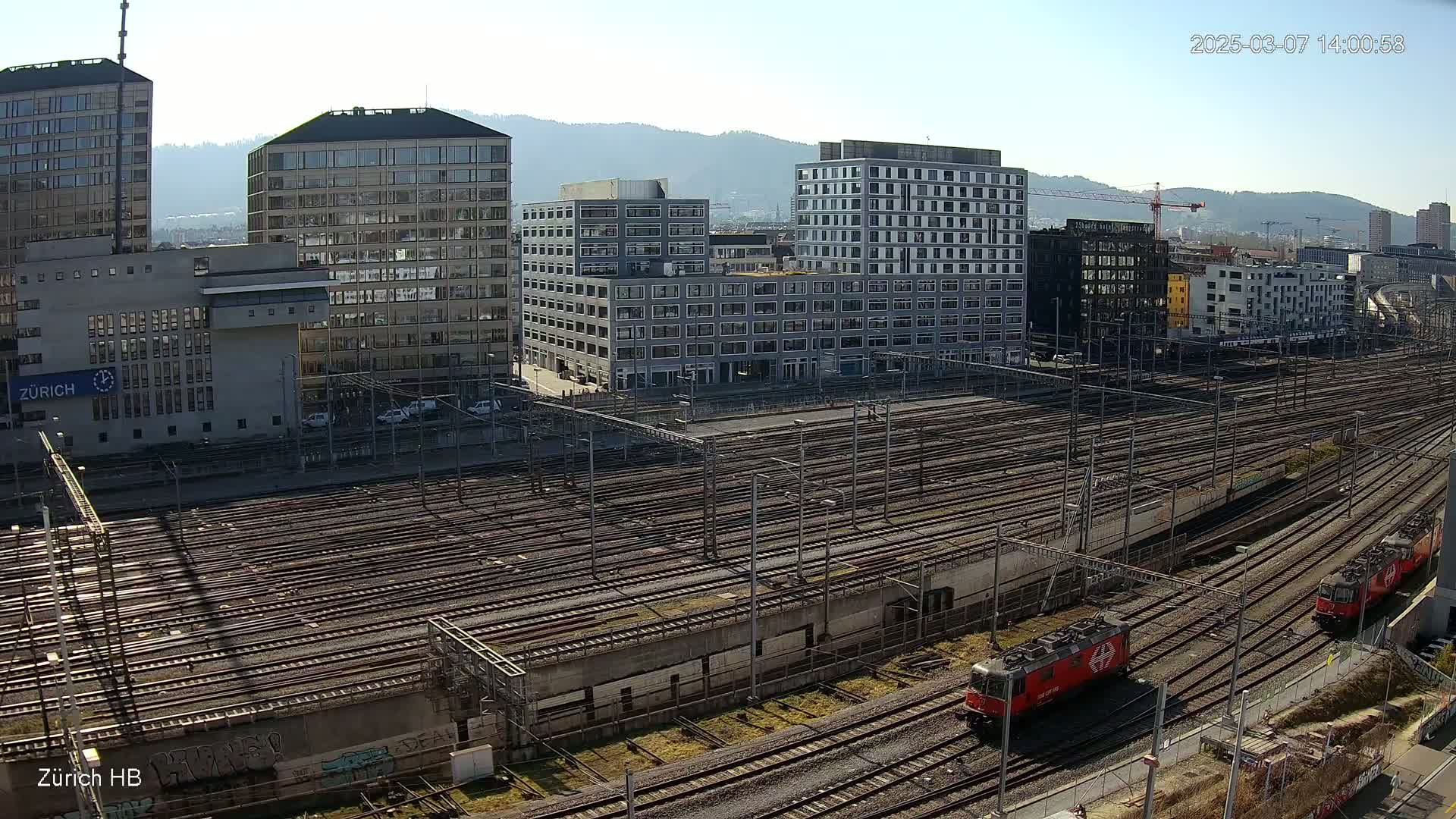 A sunny day overlooking a sprawling railway yard with numerous tracks, flanked by modern and older buildings, and two red locomotives visible in the foreground.