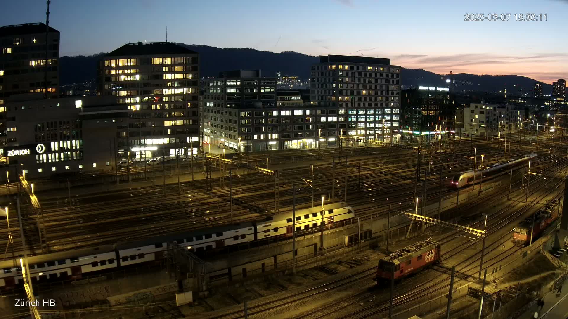 A nighttime view of a city's illuminated train station with multiple trains on numerous tracks, surrounded by buildings with lit windows under a twilight sky.