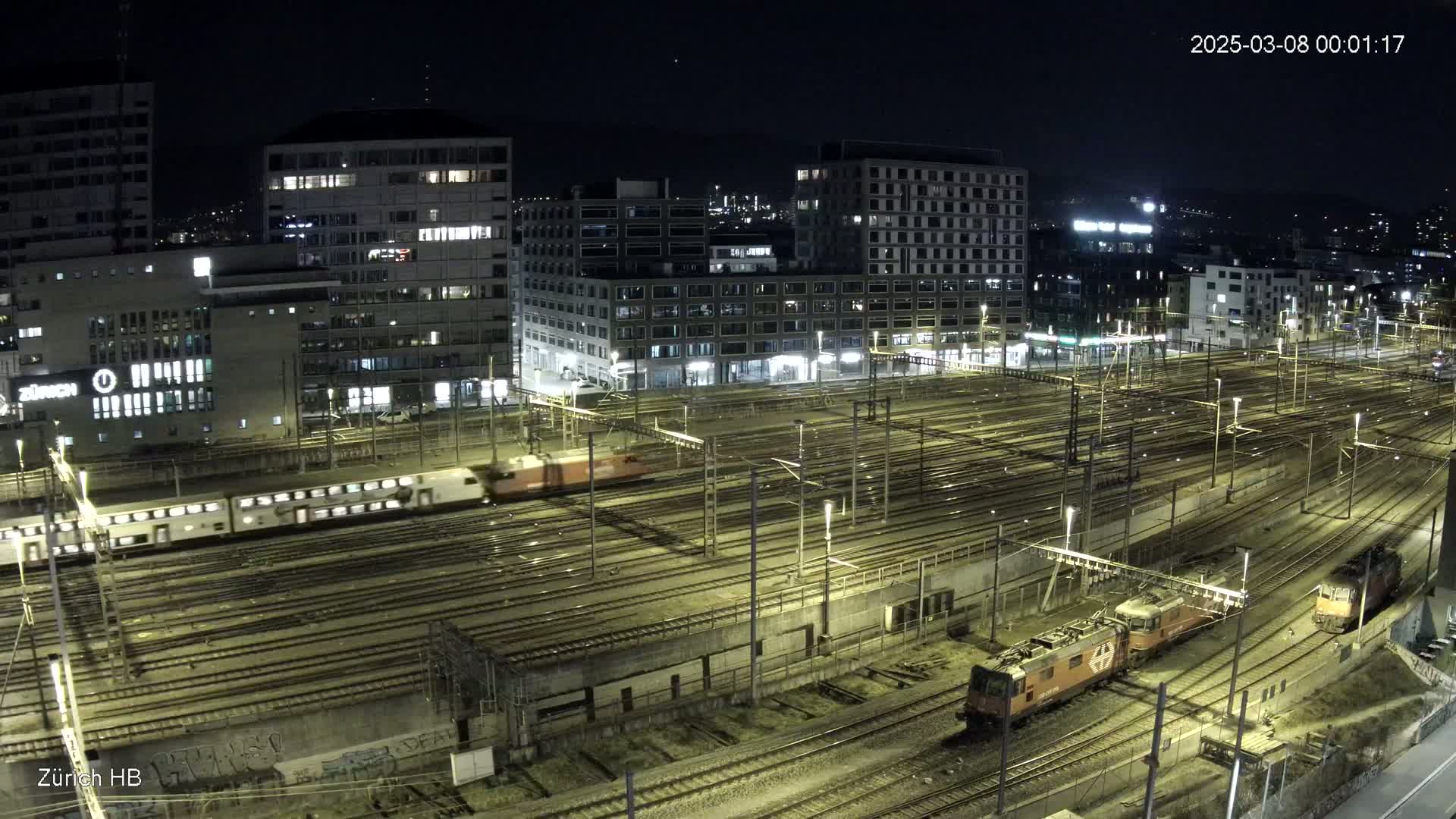 A nighttime, high-angle view of a sprawling train yard with several trains moving across numerous tracks, surrounded by illuminated city buildings.