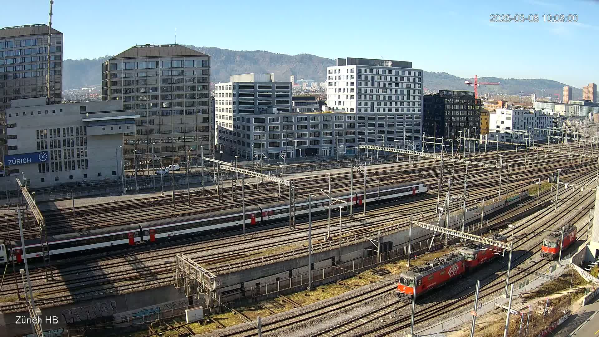 A sunny day reveals a large train yard with multiple tracks, several trains, and modern buildings in the background.