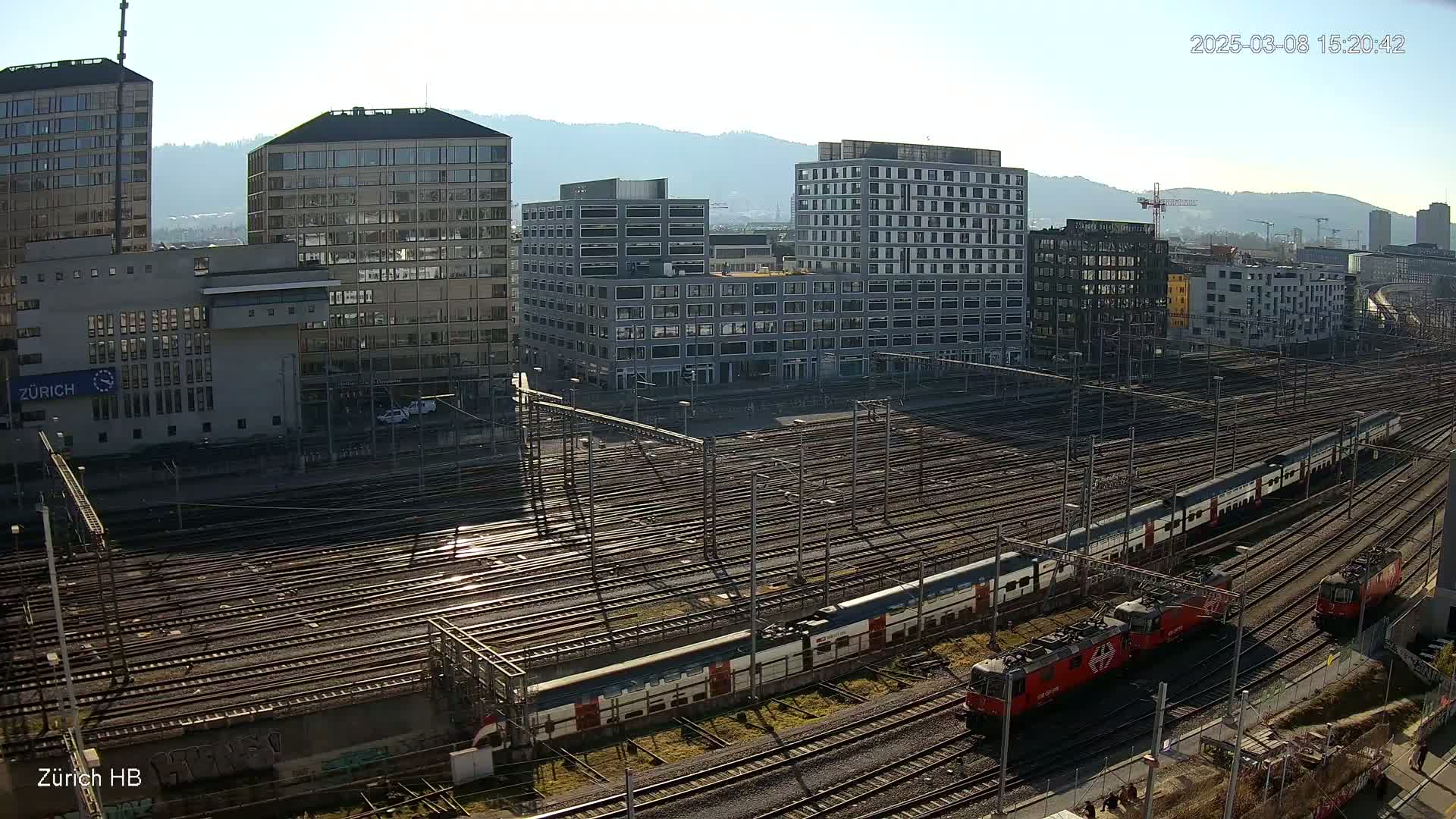 A sunny day reveals a view of a large railway yard with multiple train tracks, several modern buildings, and two passenger trains with red locomotives.