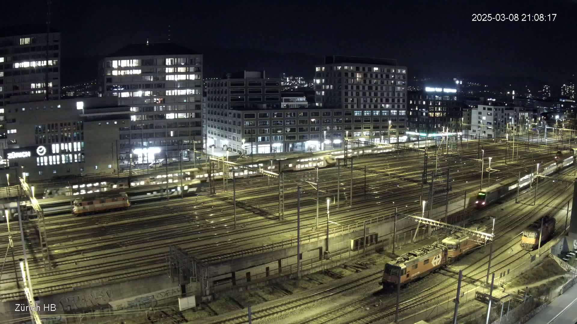 A nighttime aerial view of a bustling train yard surrounded by illuminated buildings under a clear, dark sky.