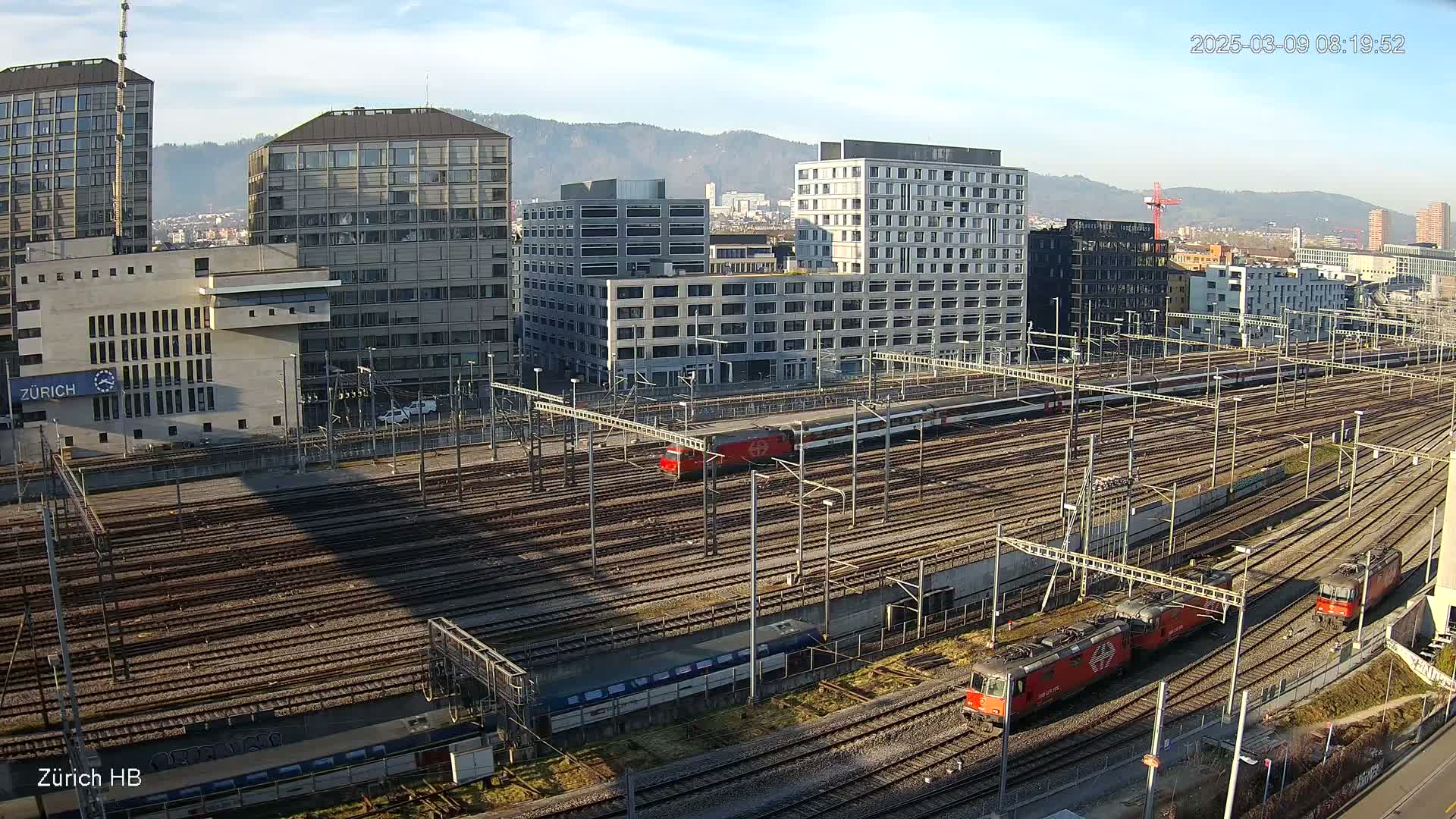 A sprawling railway yard with numerous train tracks, several trains, and modern buildings under a clear, sunny sky.