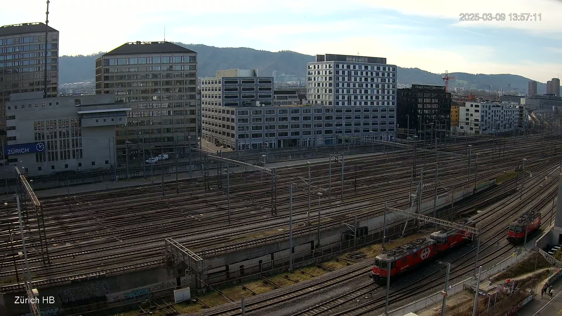 An aerial view shows a large railway yard with numerous train tracks surrounded by modern buildings under a partly cloudy sky.