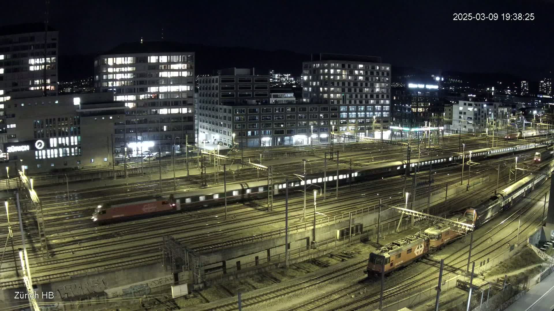 A nighttime aerial view of a busy train station with several trains on numerous tracks, surrounded by illuminated buildings.
