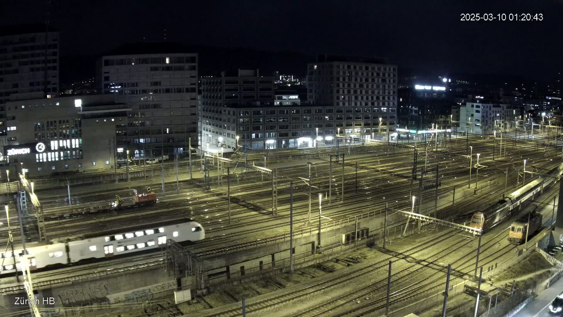 A nighttime aerial view of a large train yard with multiple trains and tracks, surrounded by buildings under a clear sky.