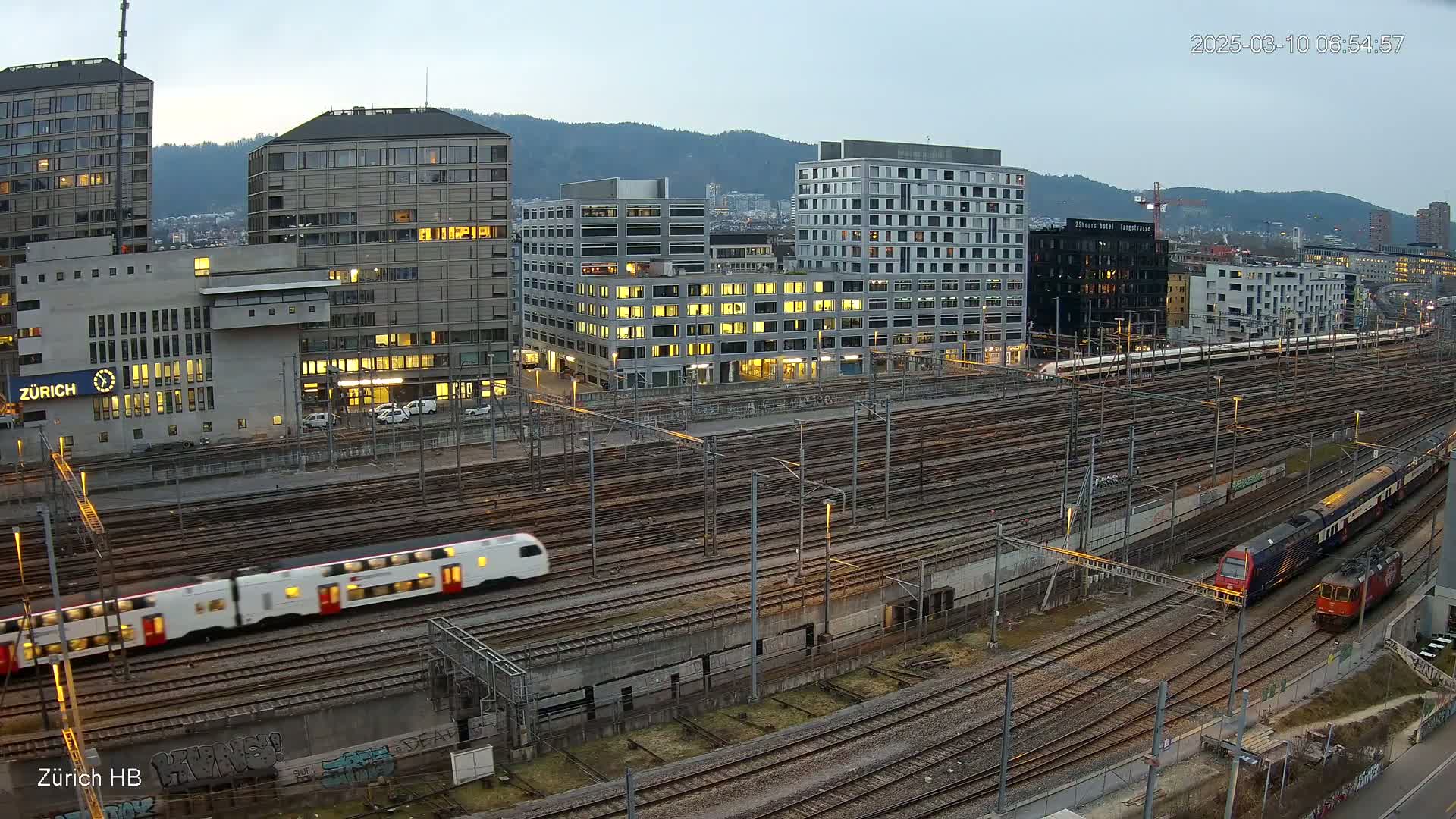 A high-angle, twilight view of a train station with multiple trains on numerous tracks, surrounded by city buildings under a partly cloudy sky.