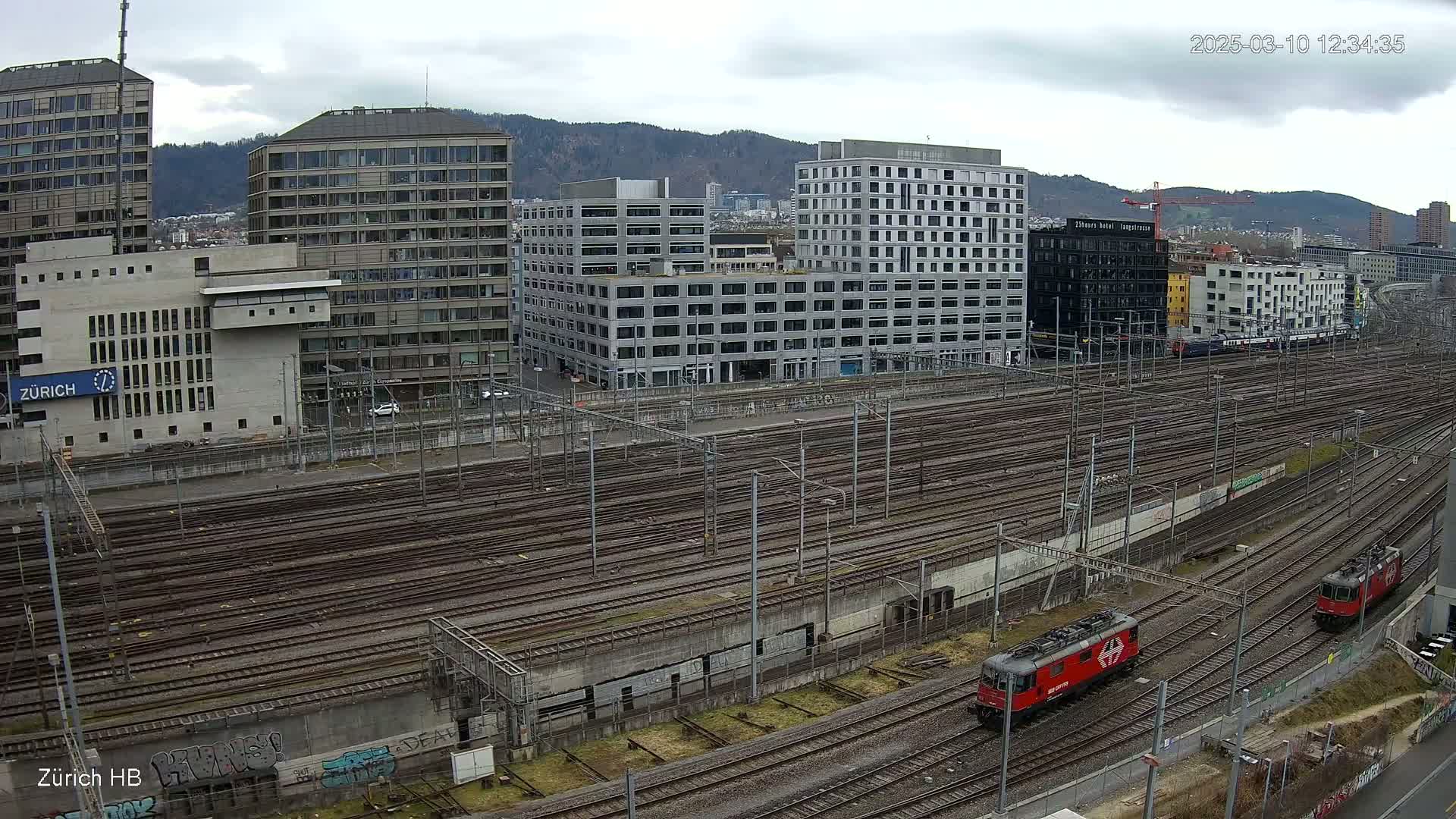 A sprawling railway yard with numerous tracks, bordered by modern buildings under a cloudy sky, features two red locomotives.