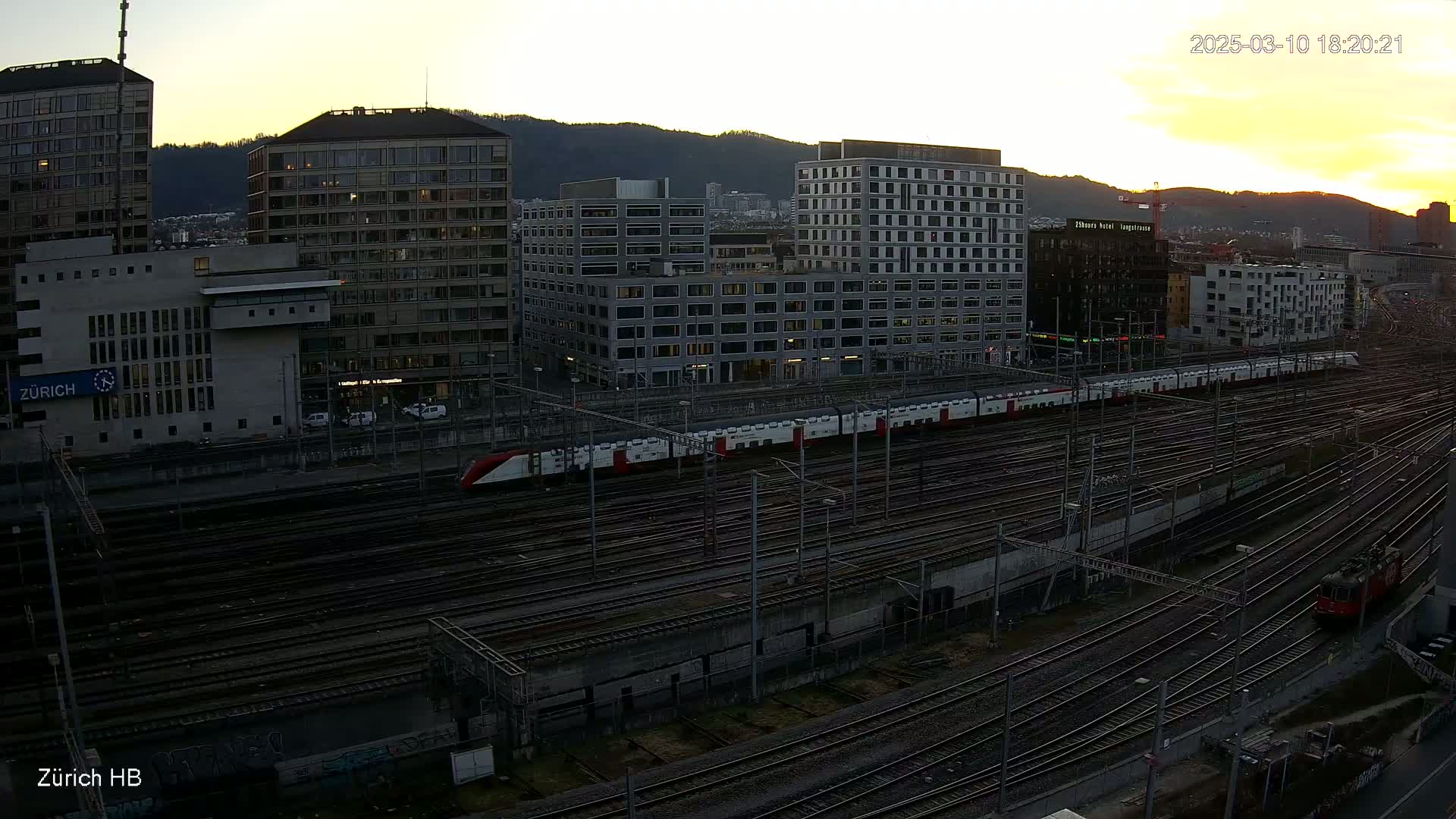 A long passenger train sits on a sprawling network of train tracks in front of several tall buildings at dusk.