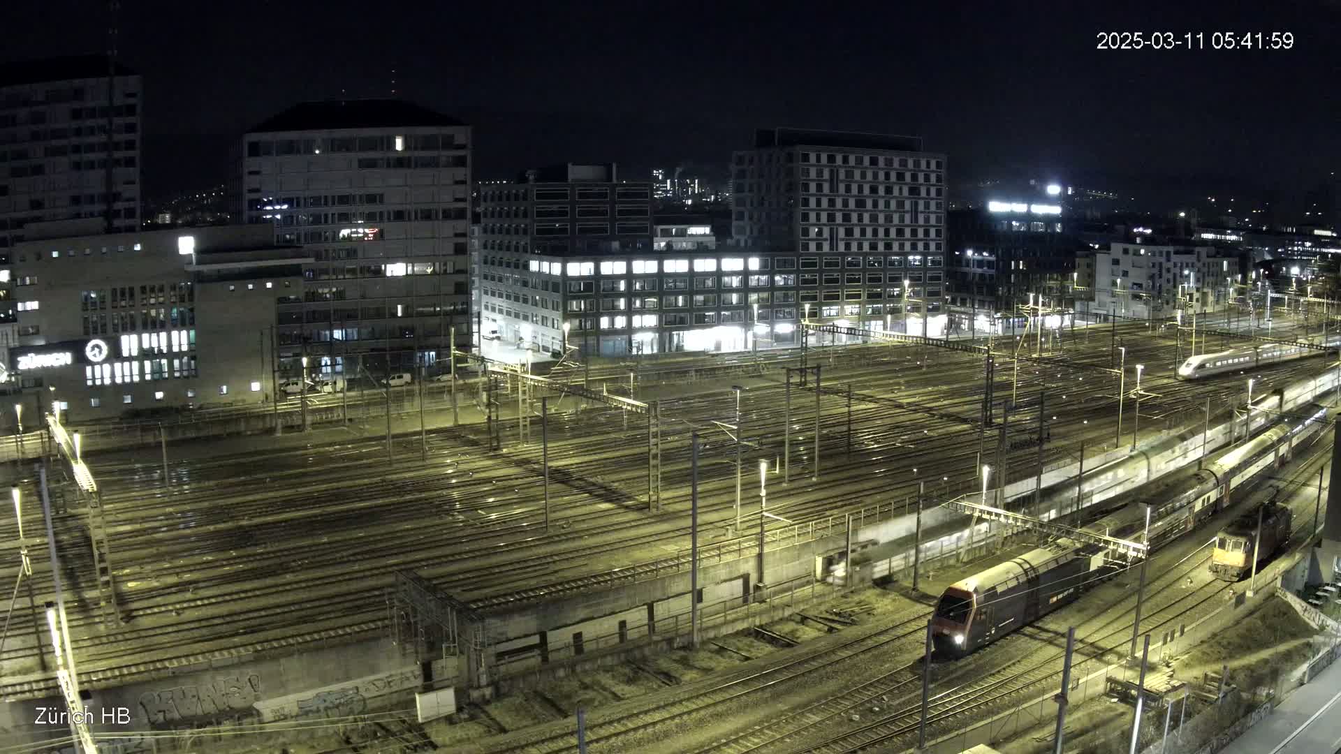 At night, two trains pass through a large, illuminated train yard surrounded by city buildings.