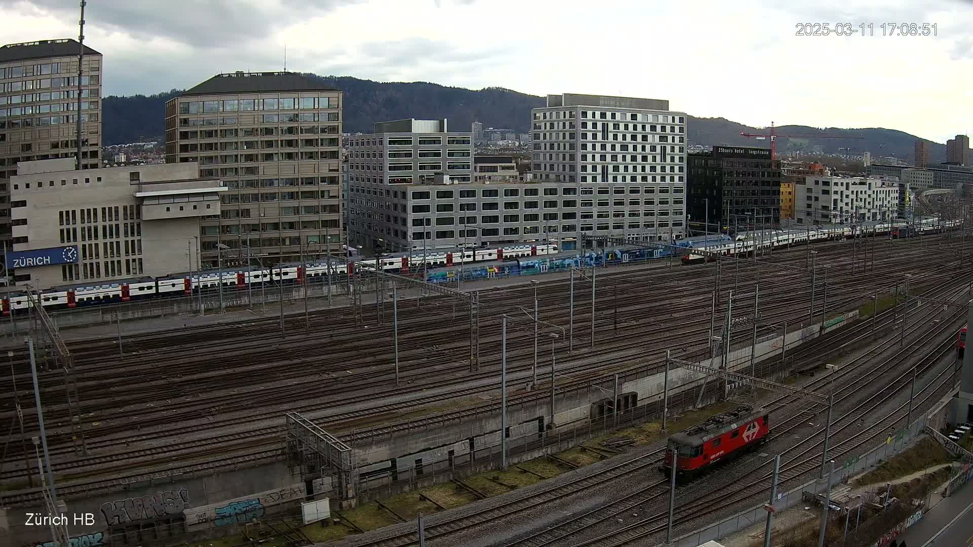 A large train station with numerous tracks, several modern buildings in the background, and a single locomotive on one of the tracks under an overcast sky.