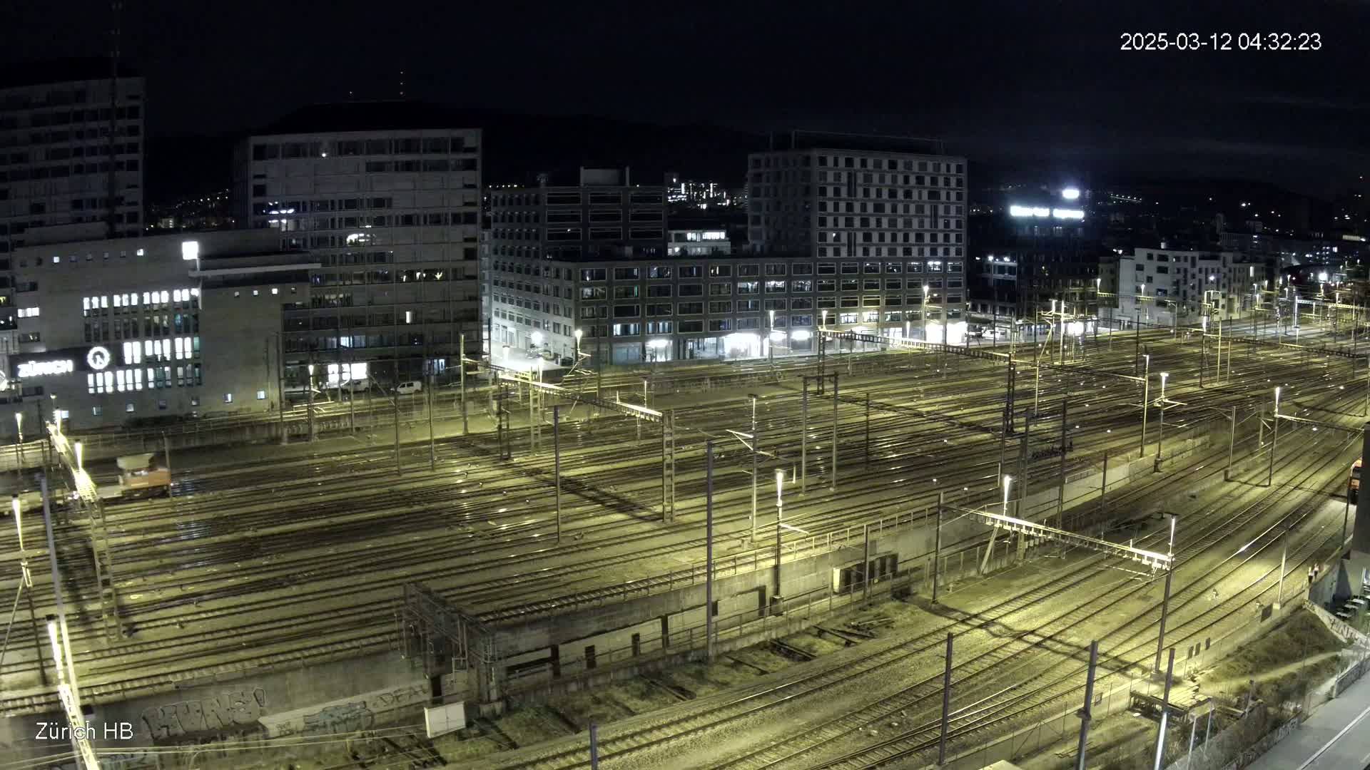 A nighttime, aerial view of a large, illuminated train yard surrounded by city buildings.