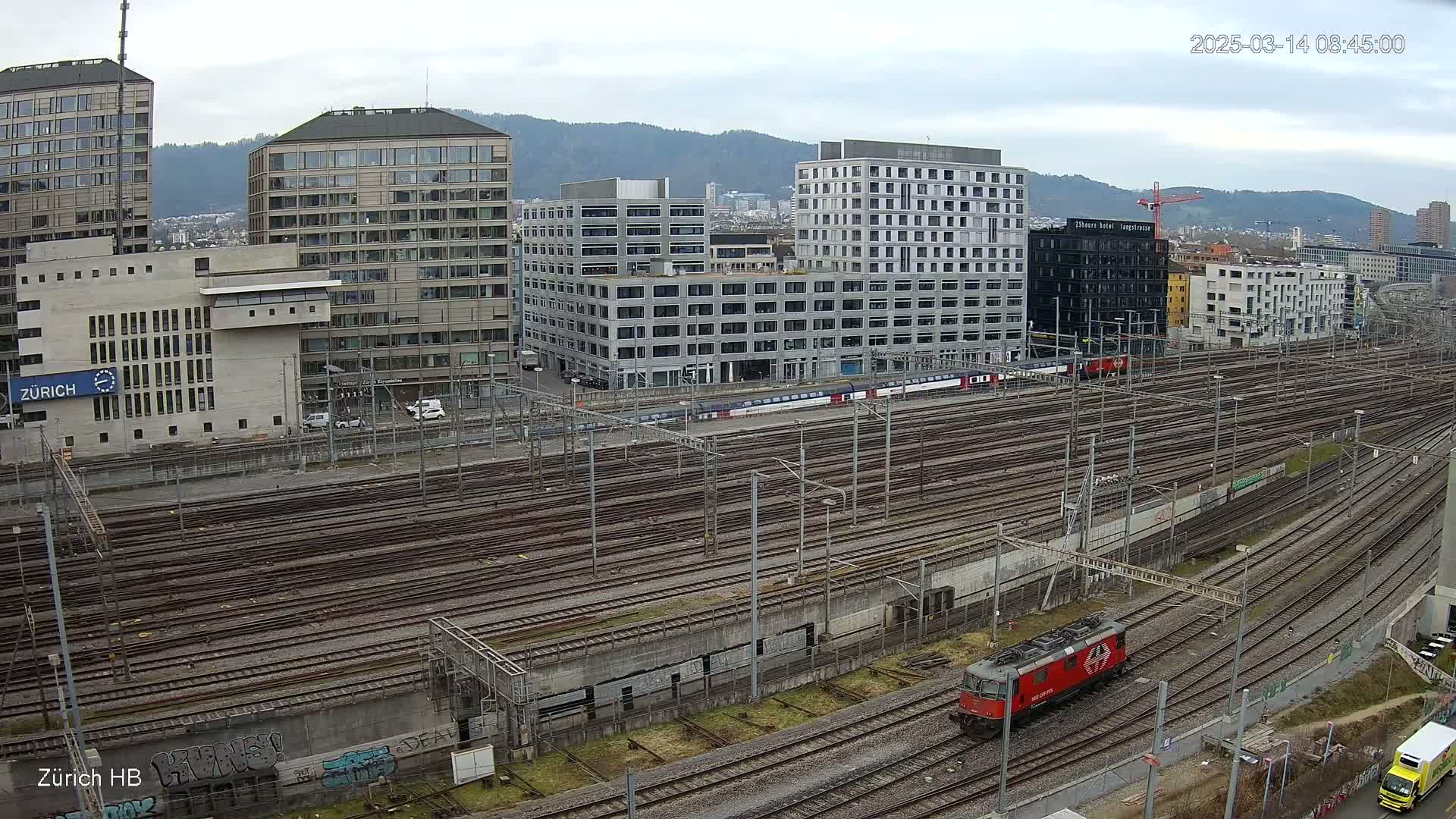 A red locomotive sits on a railway network next to several modern buildings under a cloudy sky.