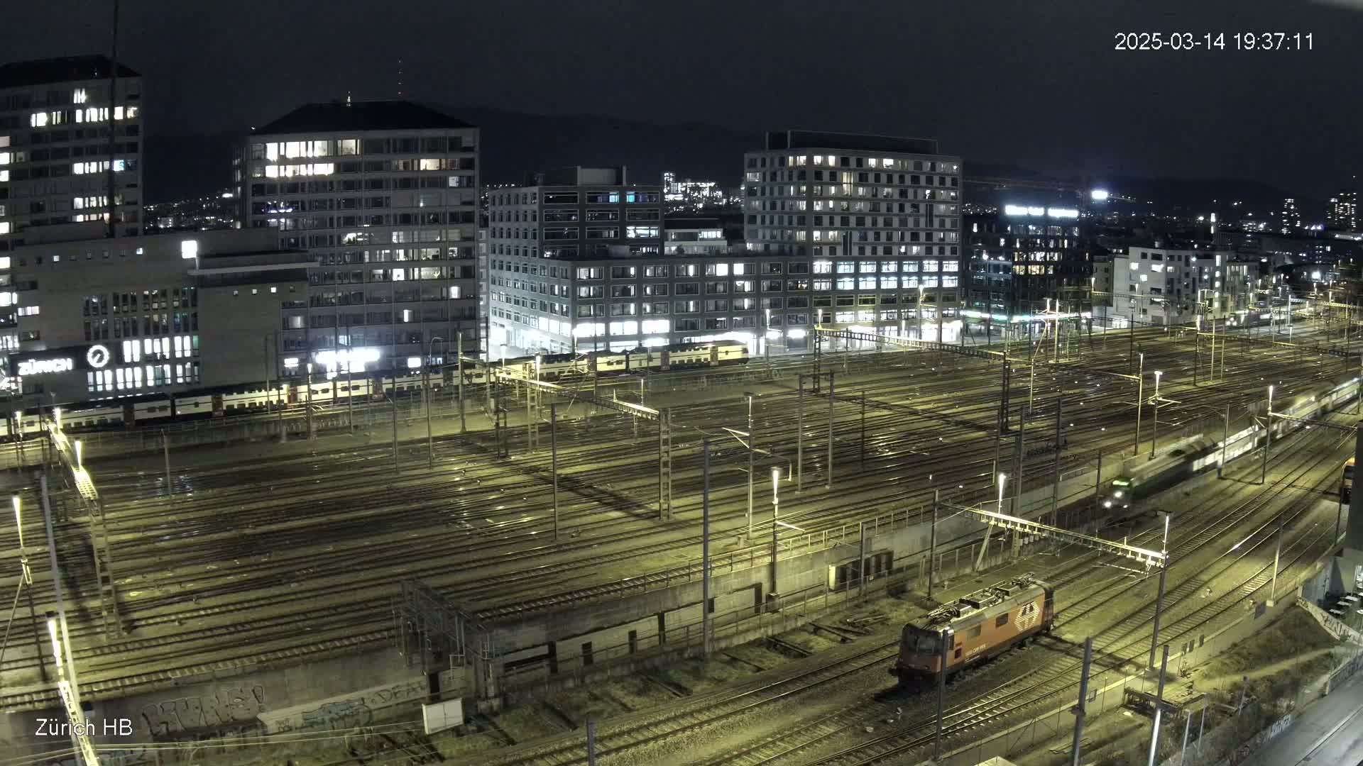 A nighttime, high-angle view of a sprawling train yard surrounded by illuminated buildings.