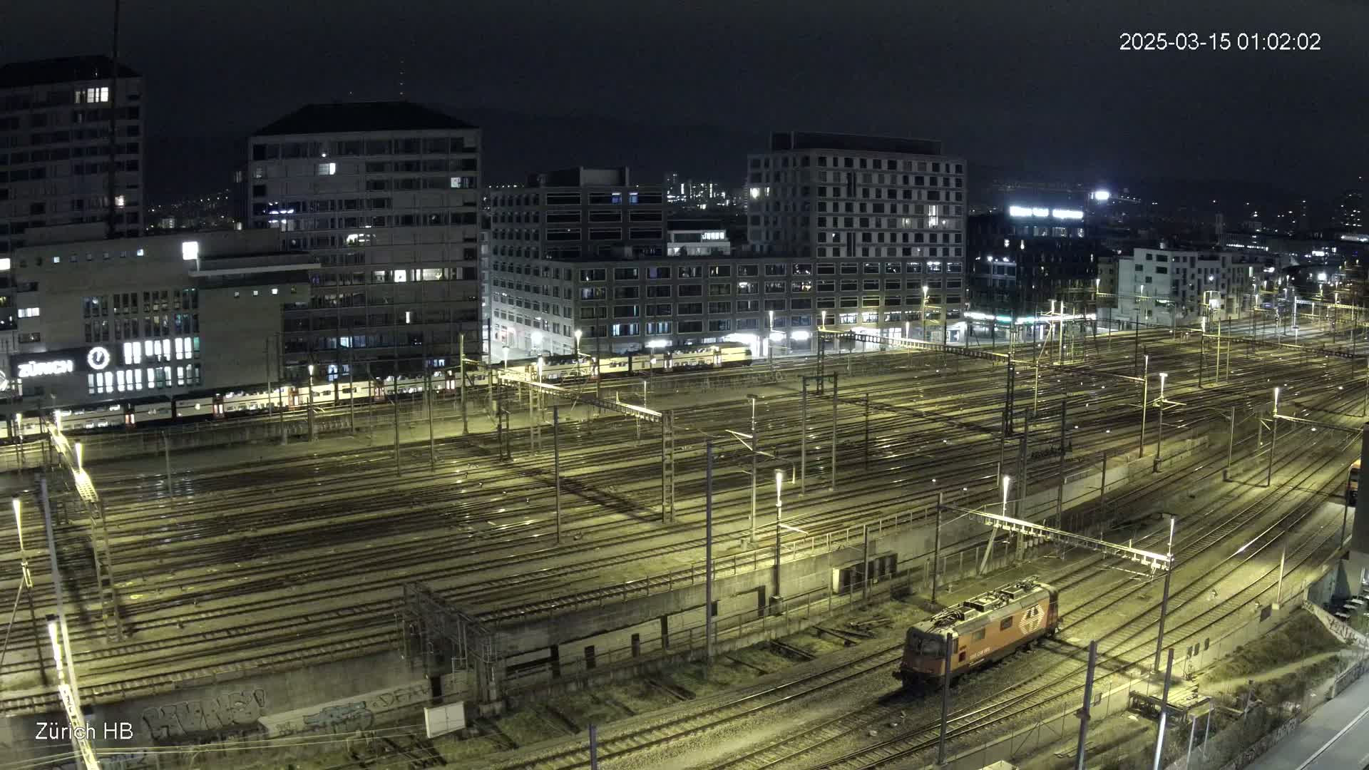 A nighttime, high-angle view of a large train yard with multiple tracks, surrounded by city buildings.