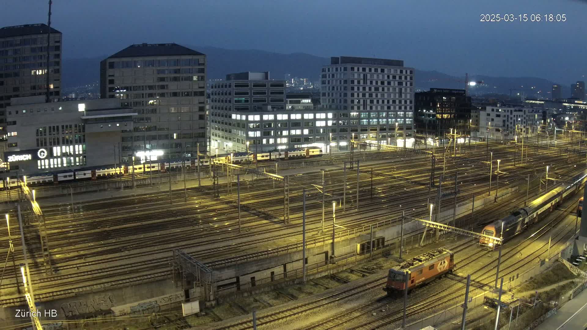A nighttime, high-angle view of a sprawling railway yard with multiple trains and illuminated buildings in the background under a dusky sky.