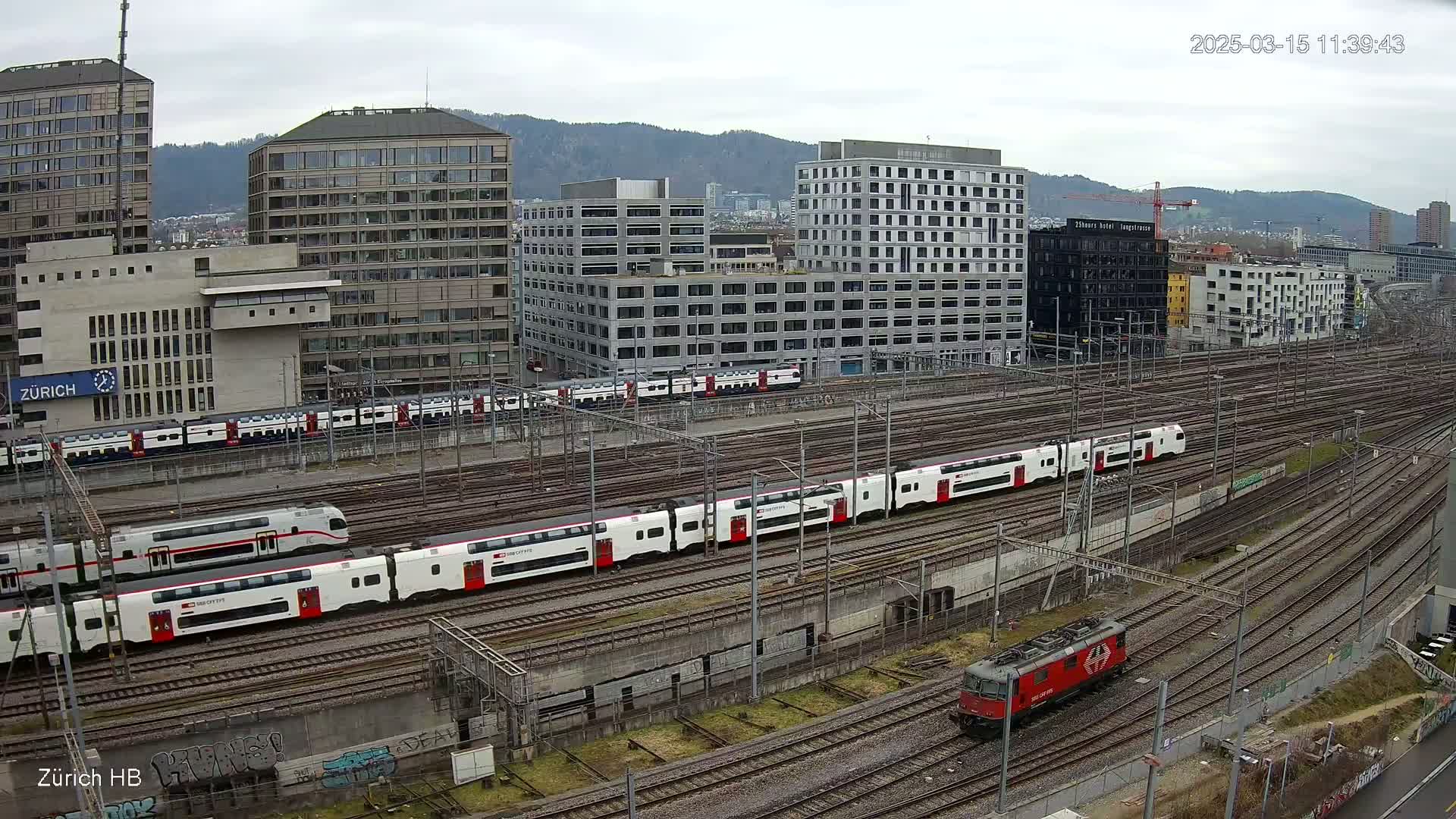 A high-angle view of a railway yard with numerous passenger trains and a locomotive under an overcast sky, surrounded by modern buildings.